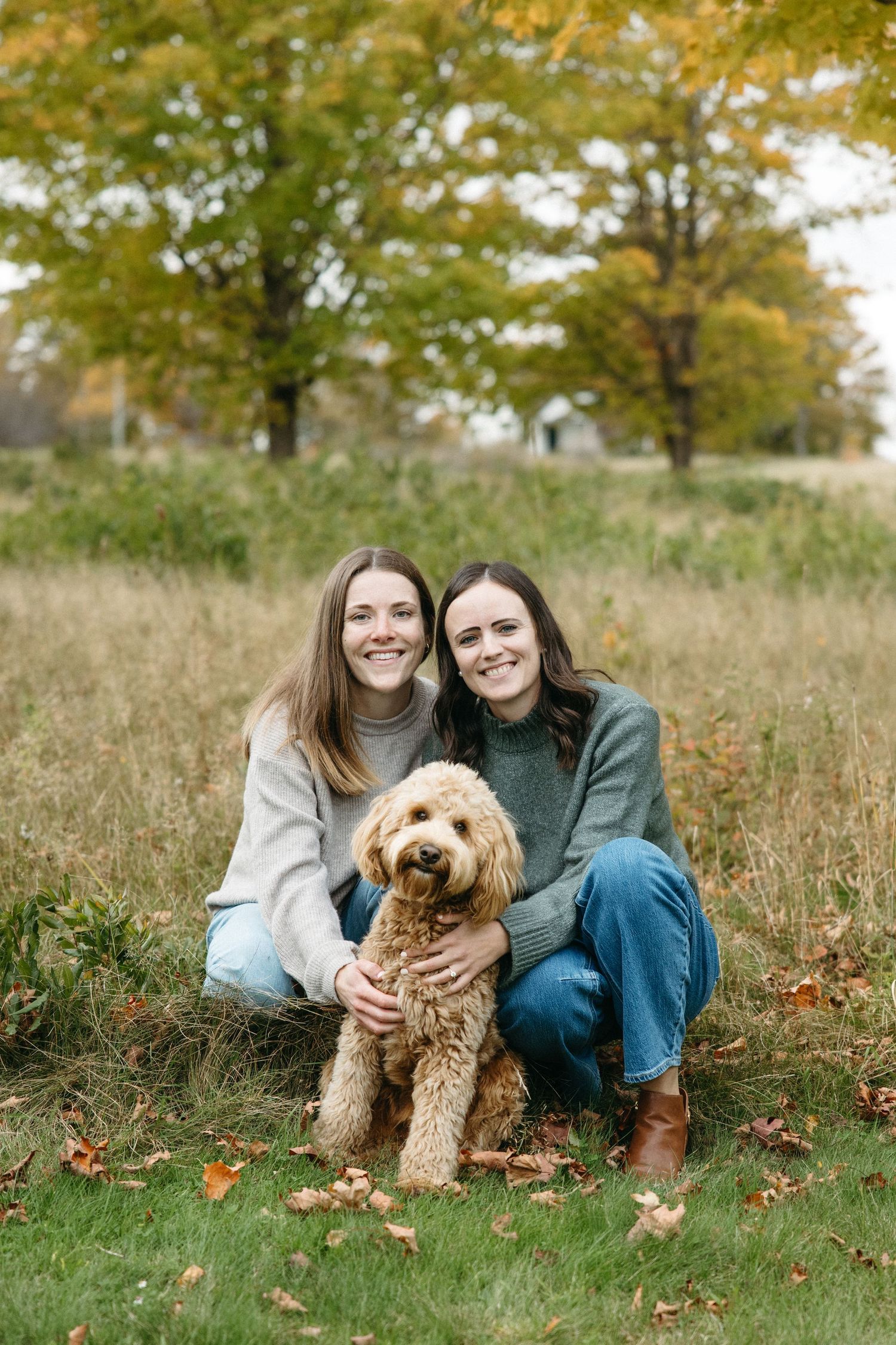 Two smiling women in sweaters sit in a grassy autumn field with a fluffy golden doodle dog between them, surrounded by fallen leaves.