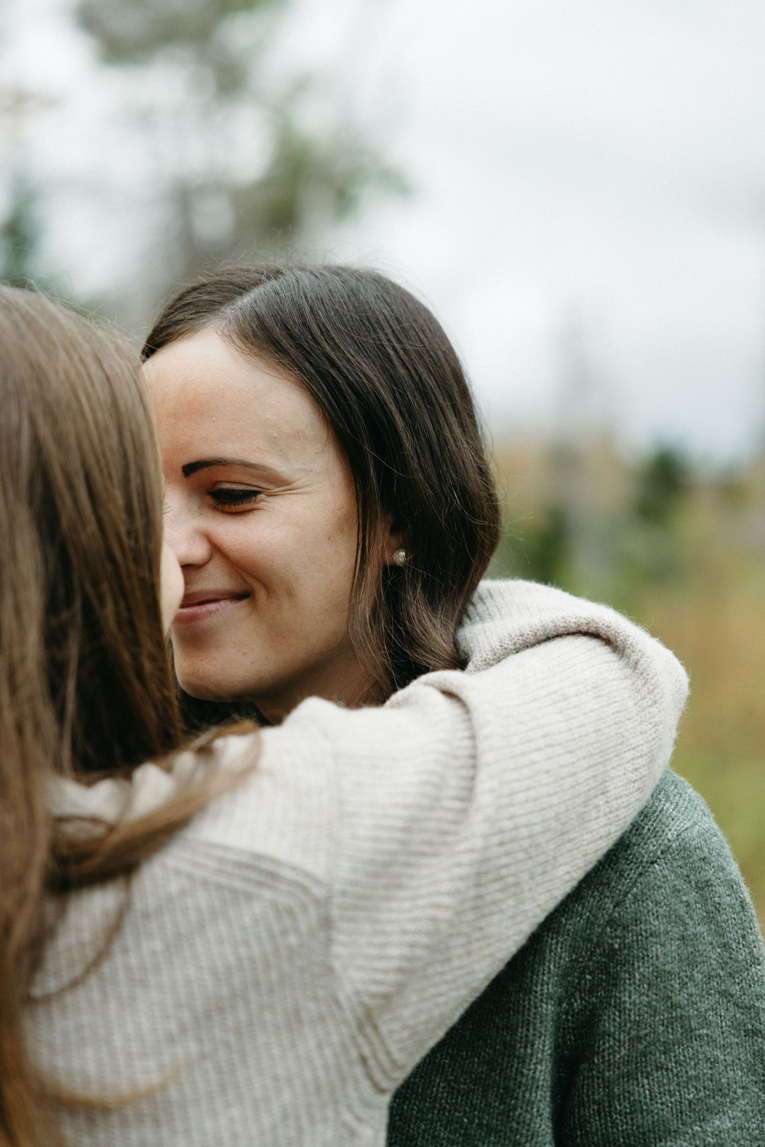 A smiling woman in a green sweater is hugged by another woman in a cream knit sweater in an affectionate outdoor portrait.