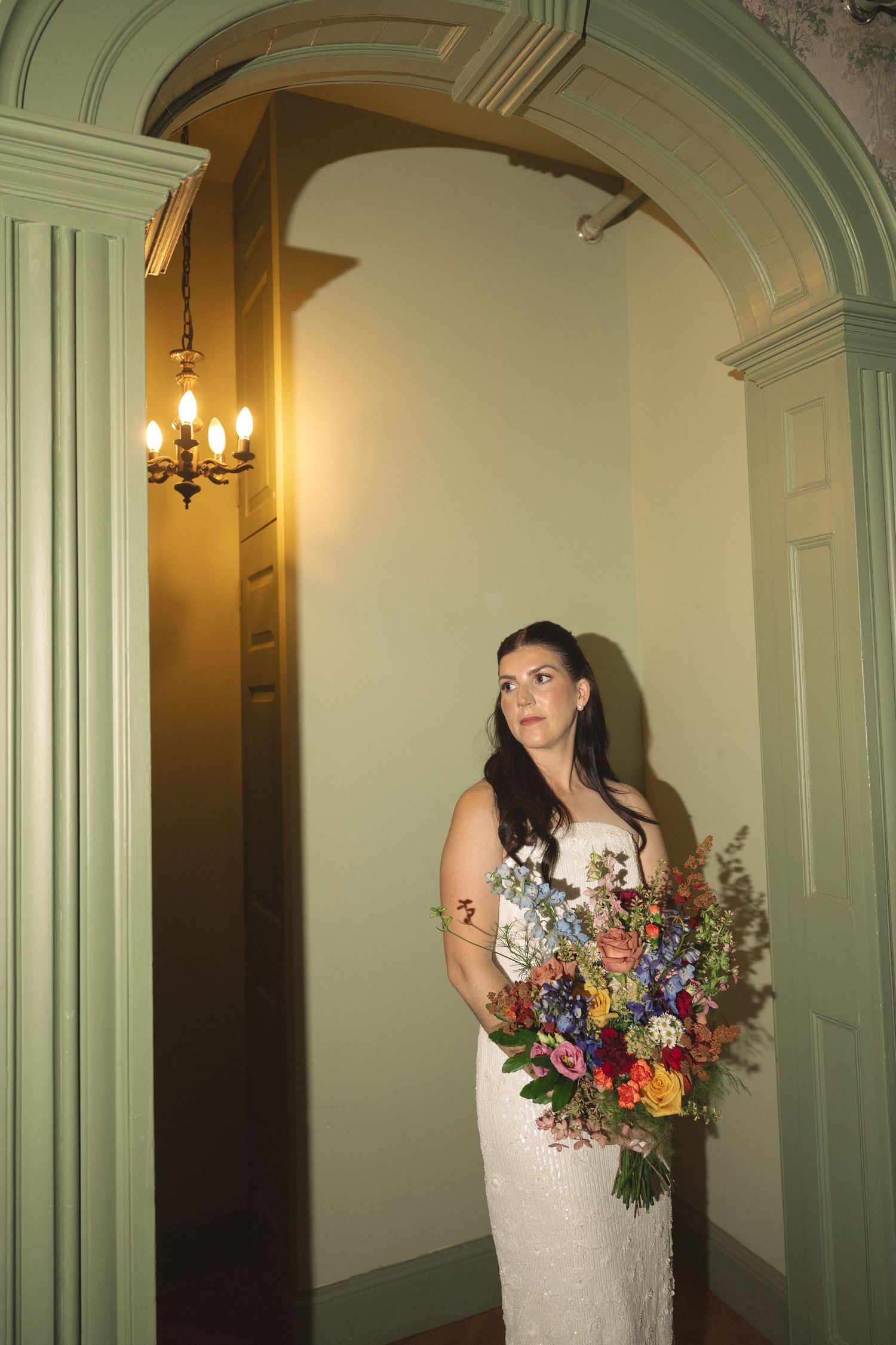 Bride holding a colorful wildflower bouquet poses in a green arched doorway beneath a warm glowing chandelier.