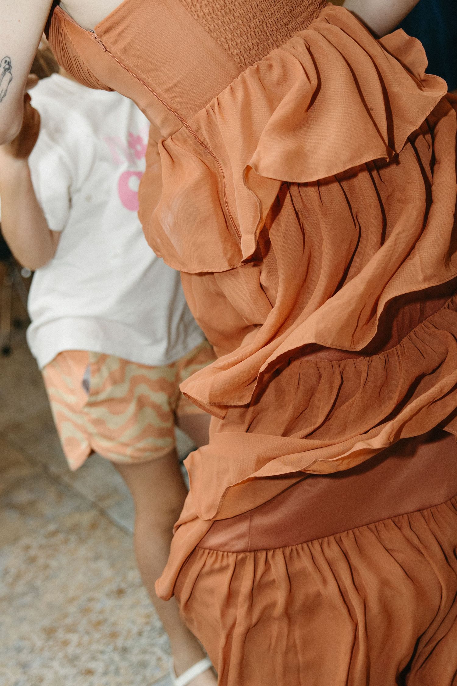 Close-up of a terracotta tiered ruffle dress in flowing chiffon beside a child in a white tee and peach patterned shorts on a tiled floor.