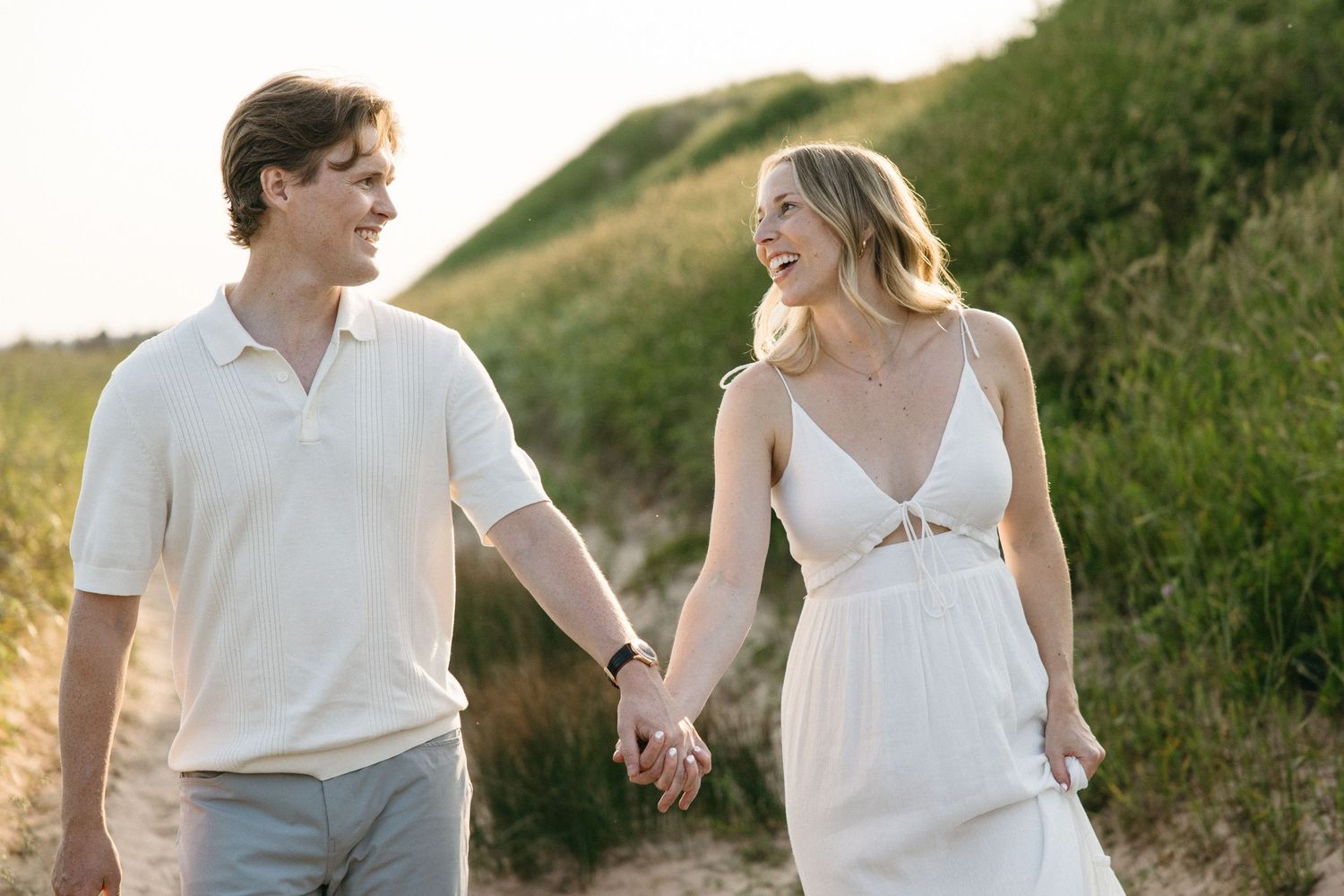 A couple holds hands walking on a sandy beach path surrounded by green dunes in golden hour light.