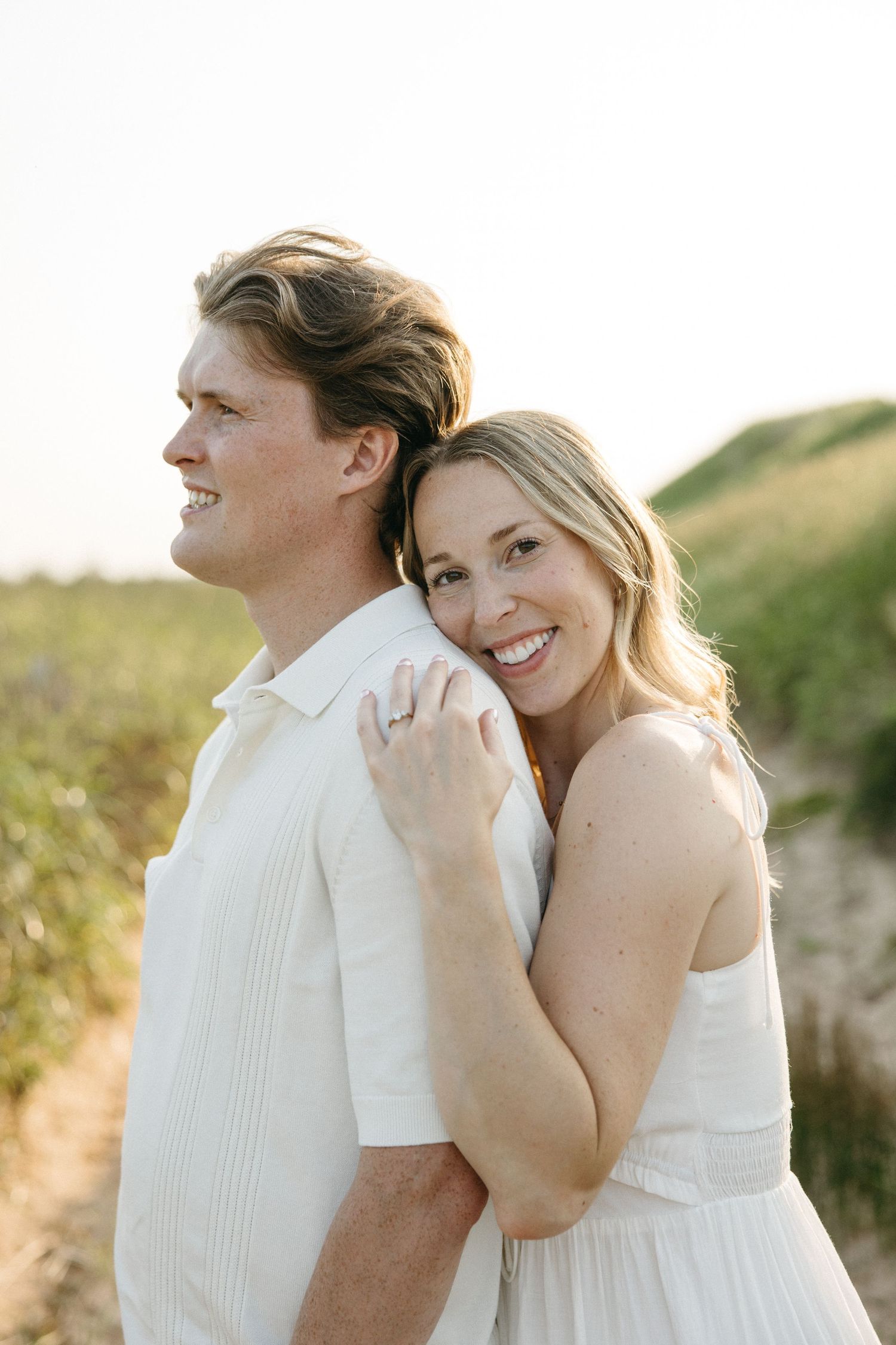 Smiling couple embracing on sandy beach path at golden hour, woman in white dress hugging man from behind.