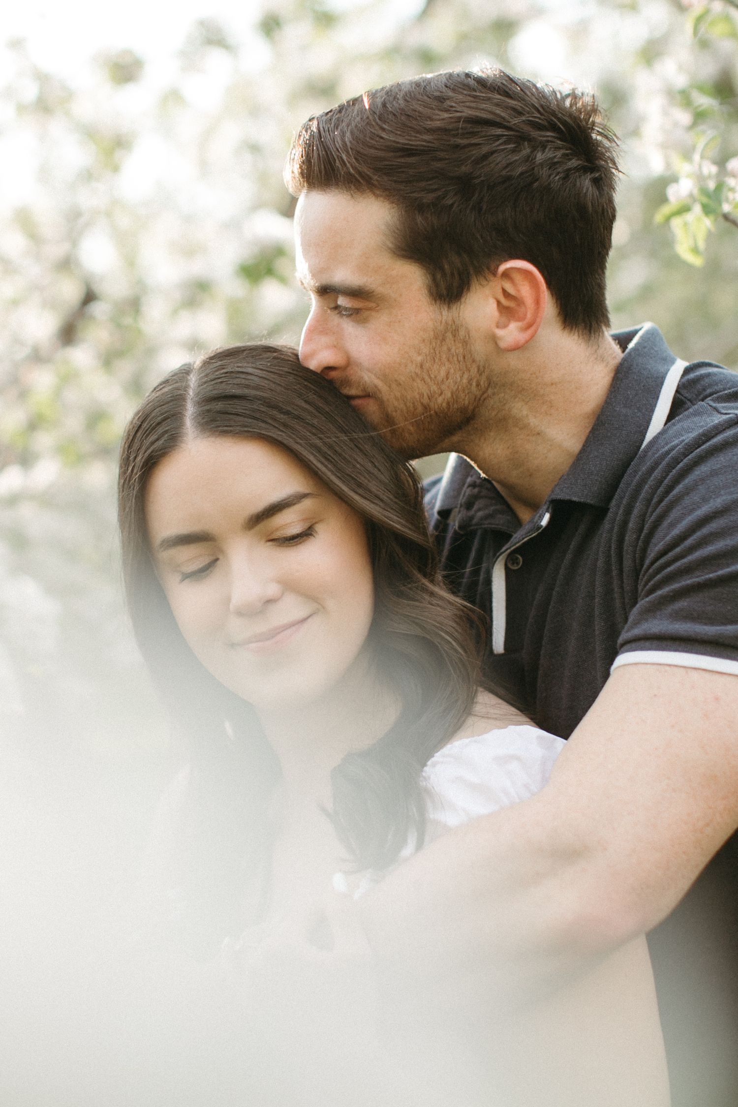 A man tenderly kisses a smiling woman on the head while embracing her outdoors near blooming white flowers.
