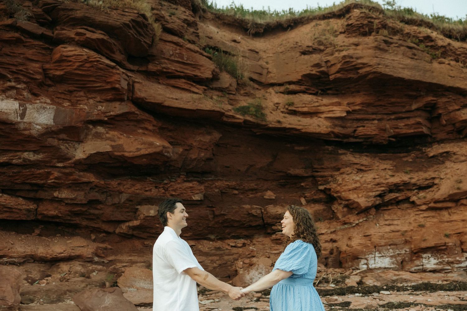 A couple shares a romantic moment on a beach with dramatic red cliffs as their backdrop.