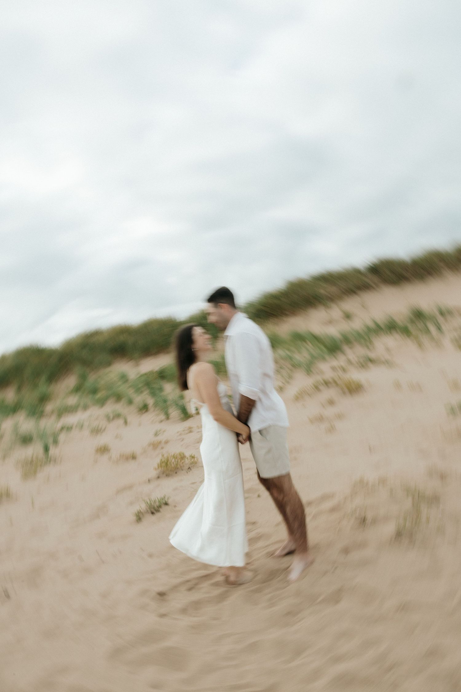 Two people in white clothing share tender moments on windswept coastal dunes.