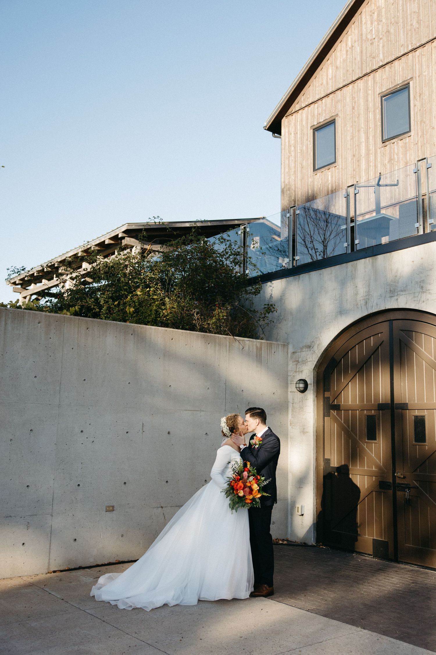 A couple shares an intimate moment in front of a modern concrete and wood building entrance.