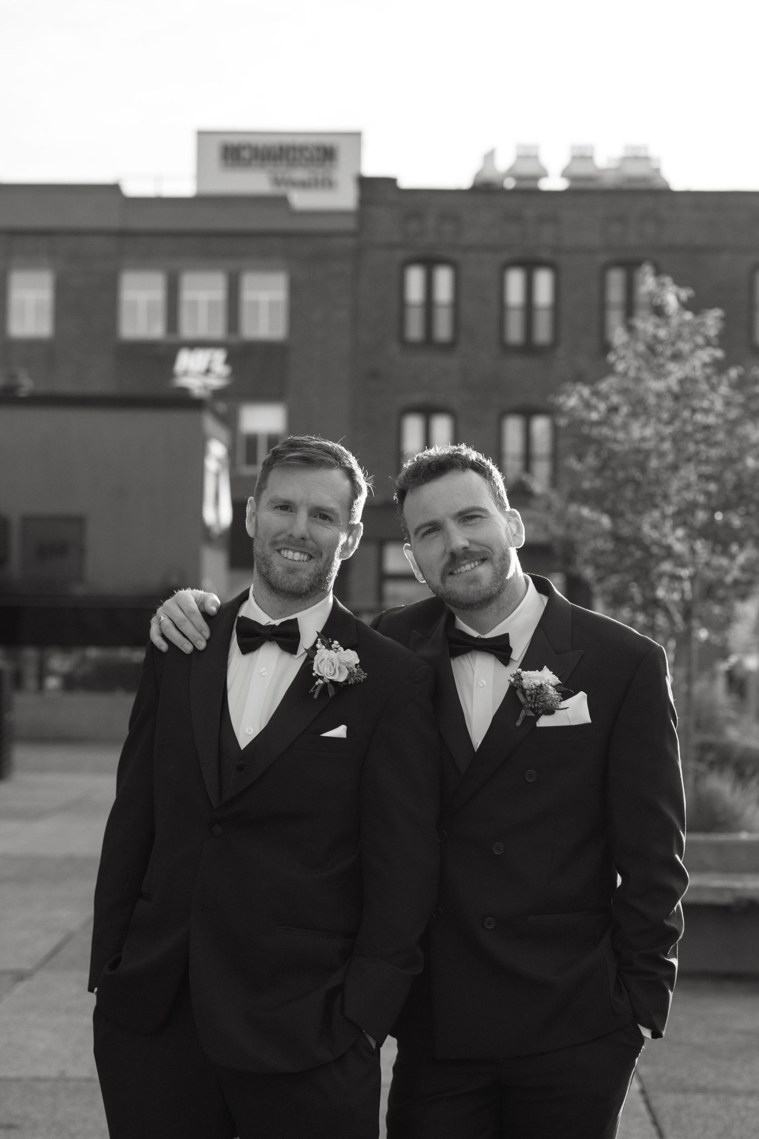 Wedding party members in black tuxedos pose together outdoors in black and white photography.