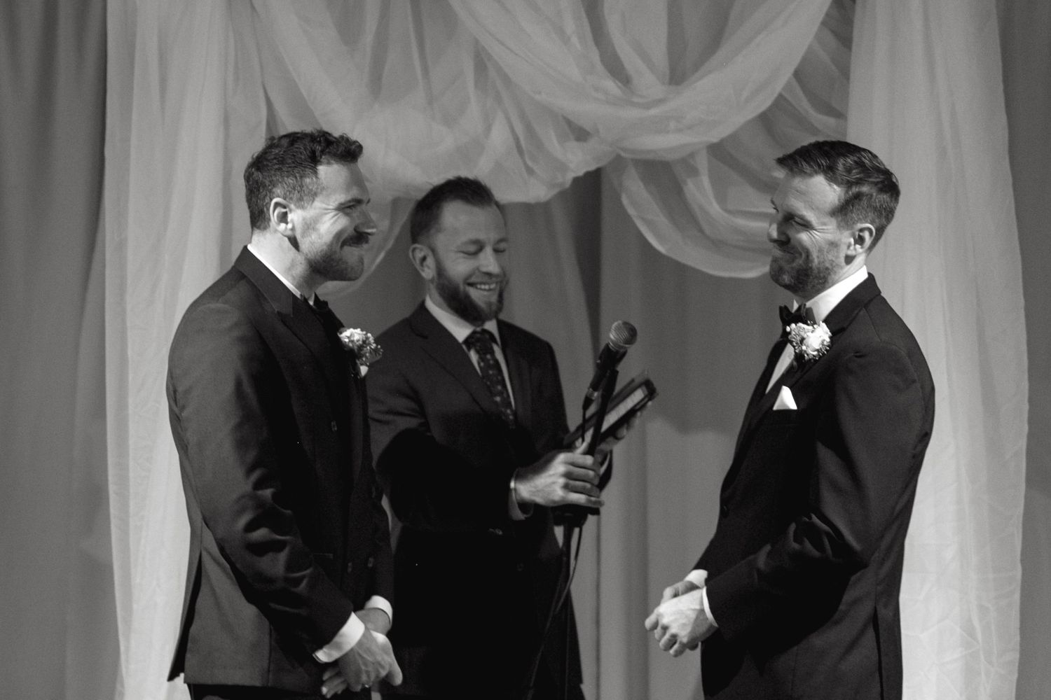 A black and white ceremony moment with three people in formal attire against draped fabric.