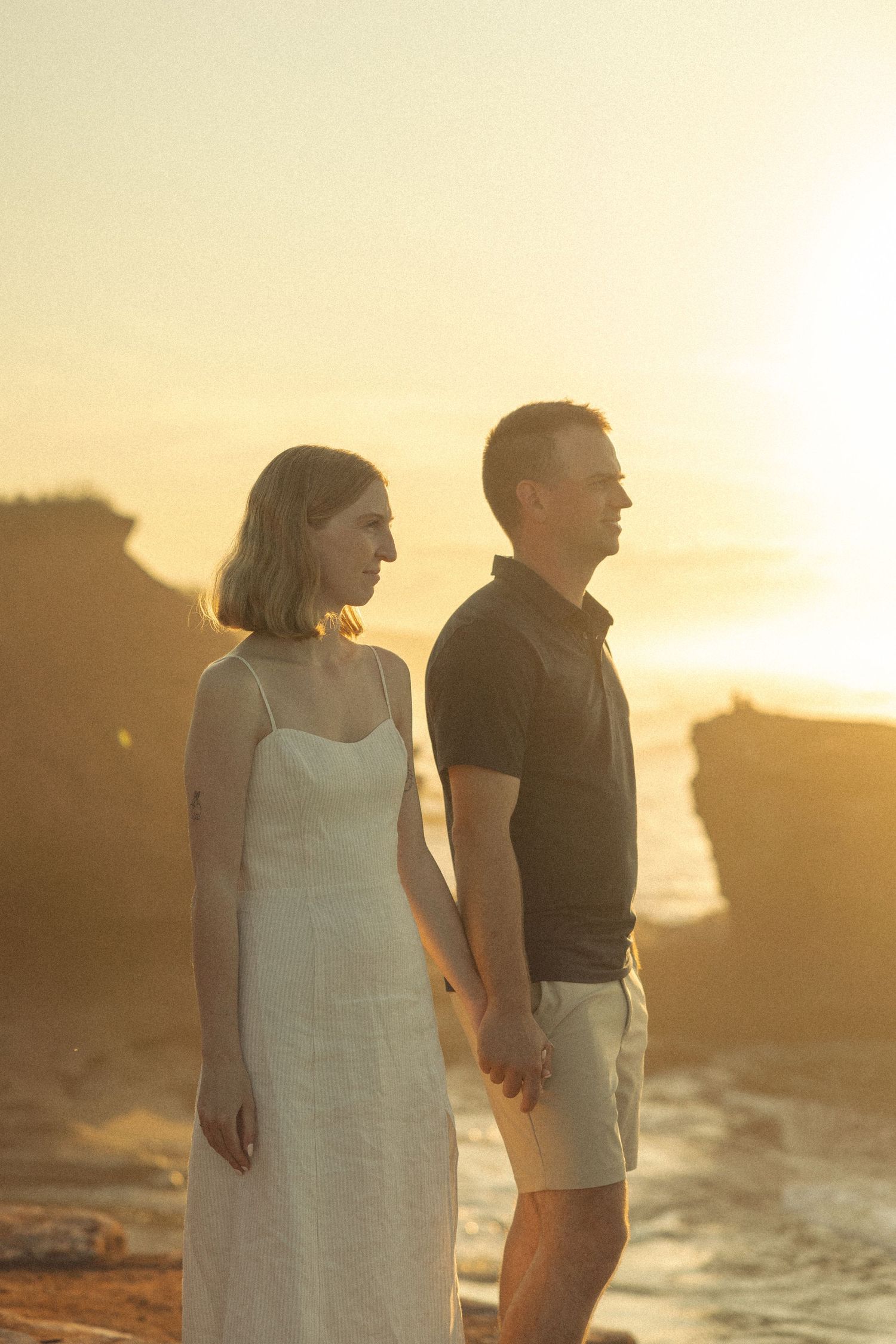 Couple in casual attire stands together on coastal cliffs during golden hour.