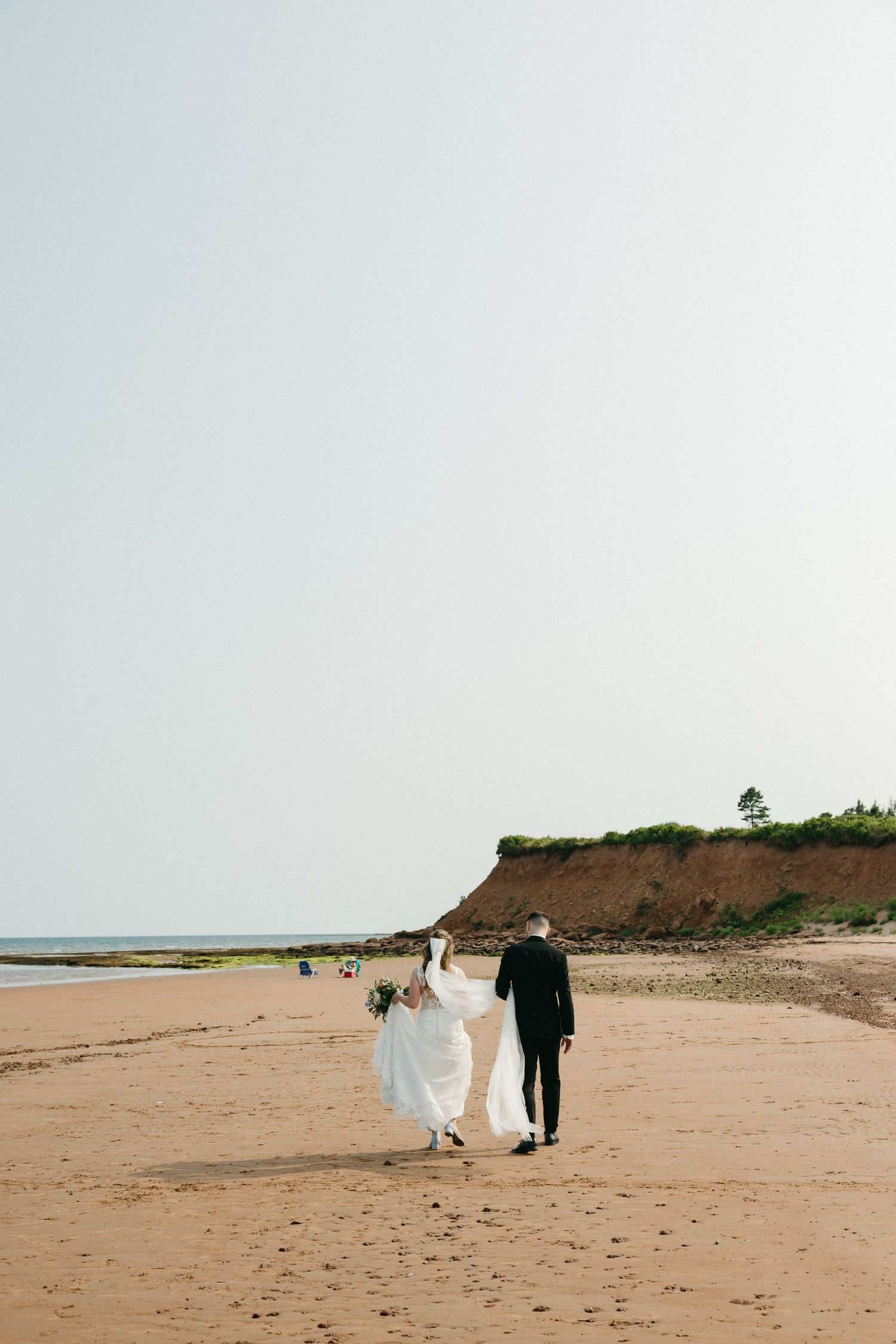 Distant view of wedding couple walking on a serene beach with cliffs in background.
