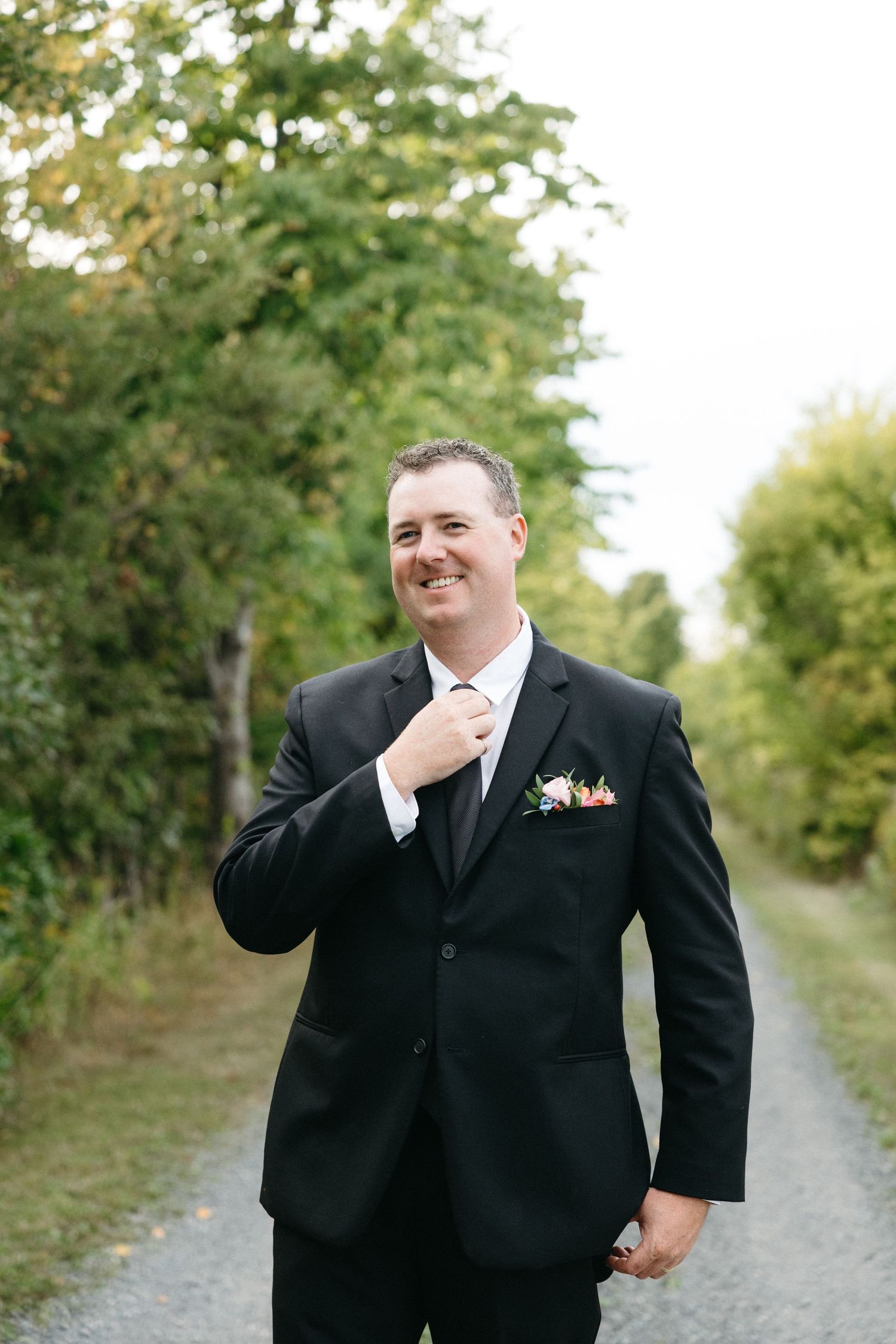 Person in black suit walking along a tree-lined gravel path.