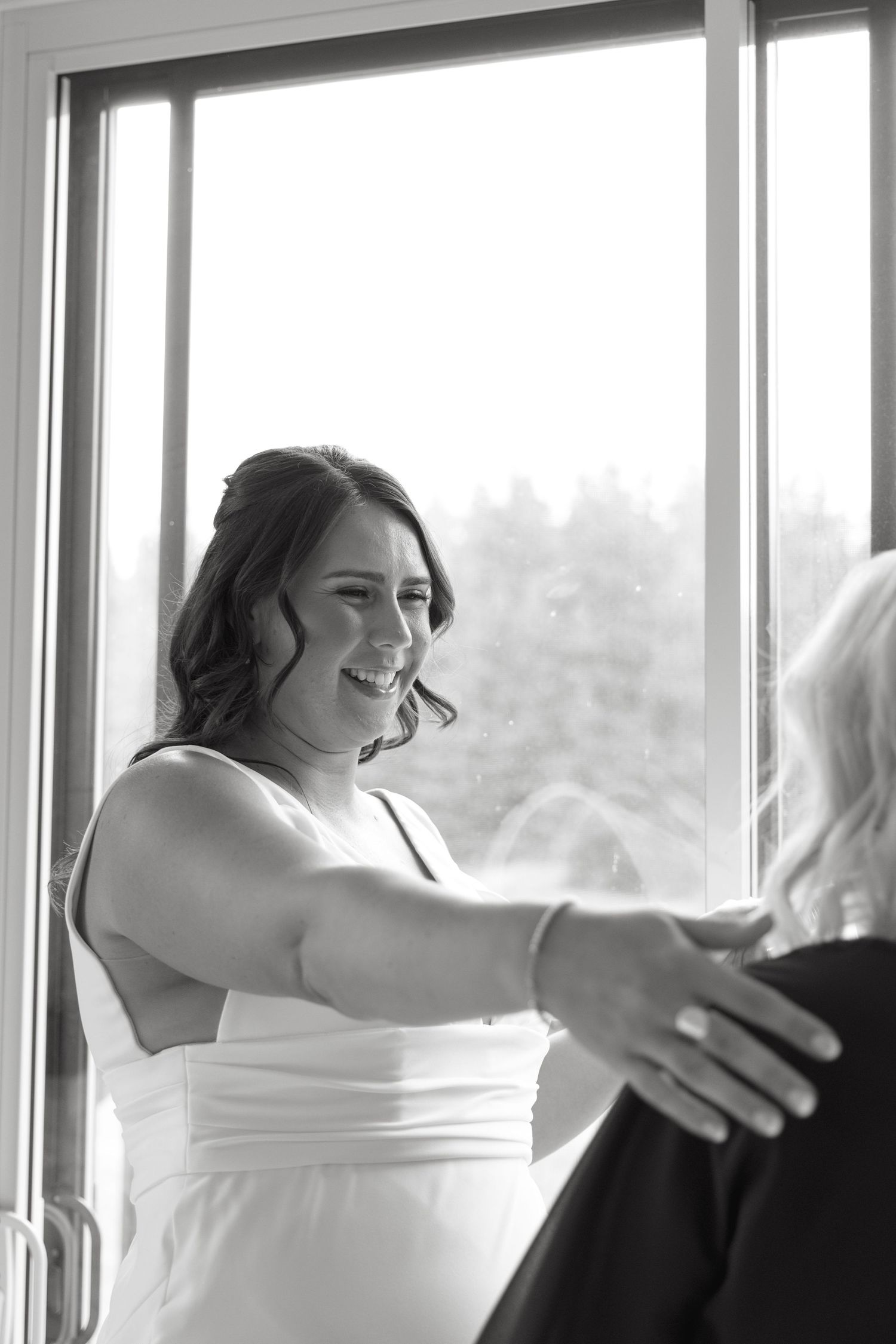 Wedding party members share joyful embraces by large windows in a bright indoor space.
