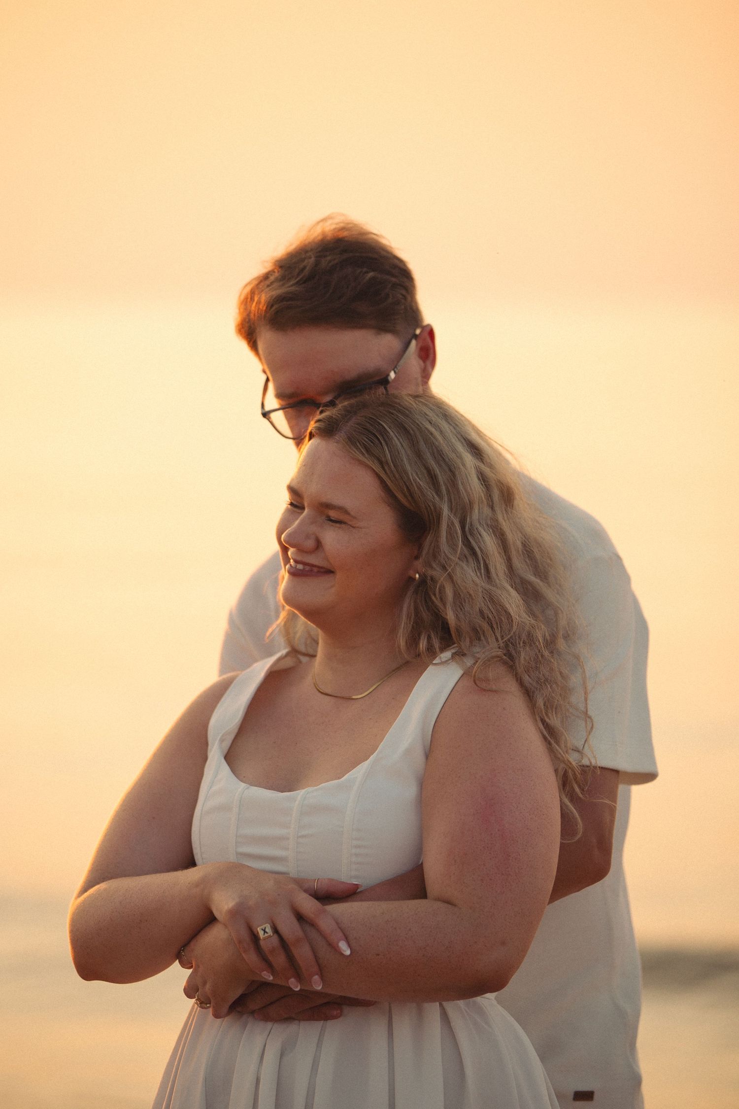 Couple embracing at golden sunset on beach, woman in white dress smiling, man holding her from behind.