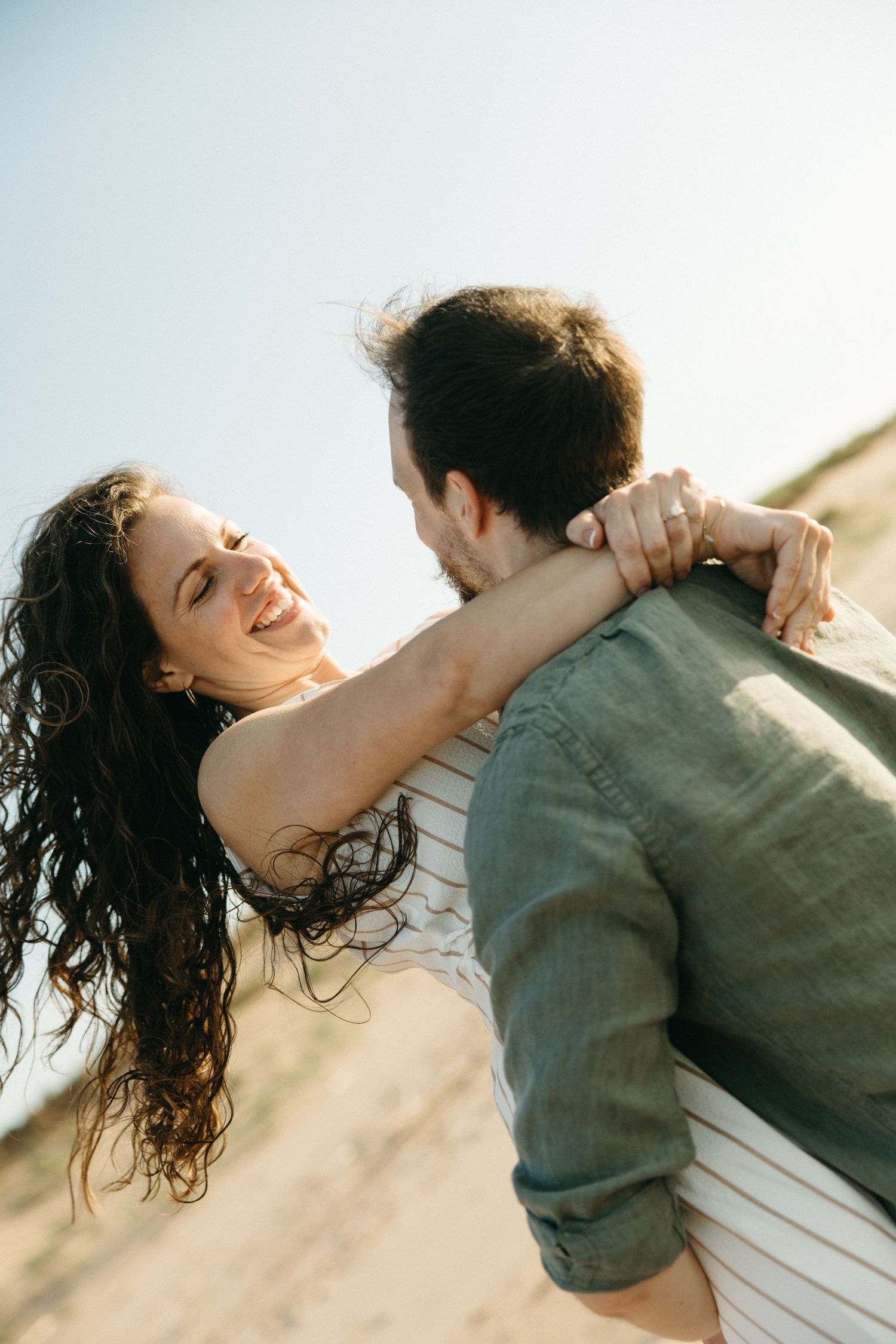 Happy couple embracing outdoors, woman laughing joyfully as man lifts her against a sunny blue sky.