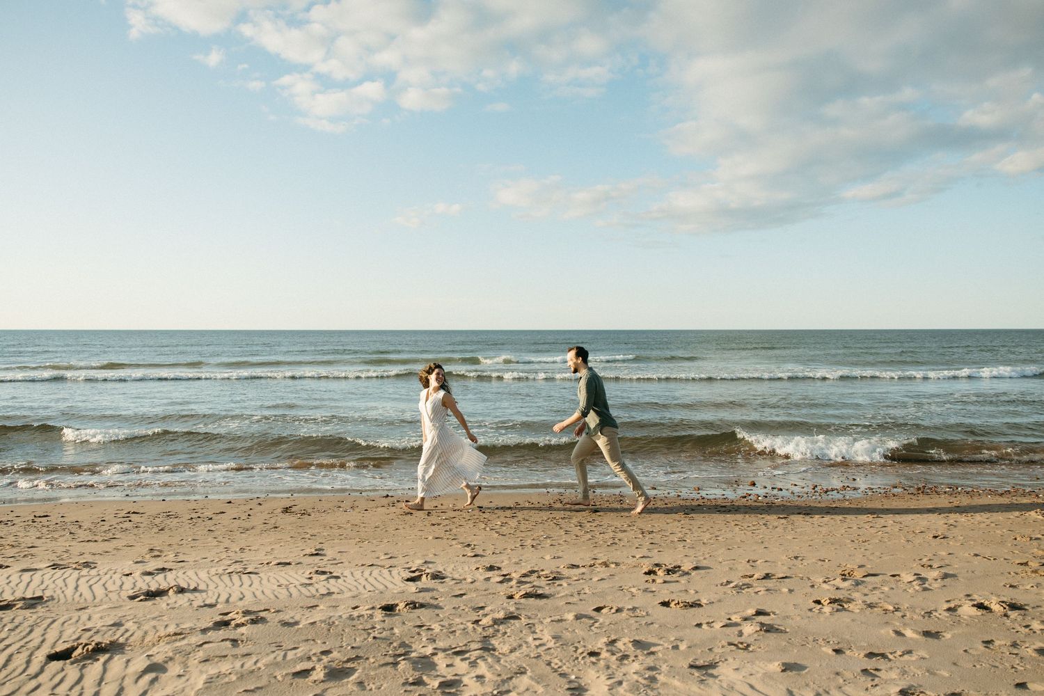 Couple running hand in hand along sandy beach shore with blue sky and ocean waves in background.