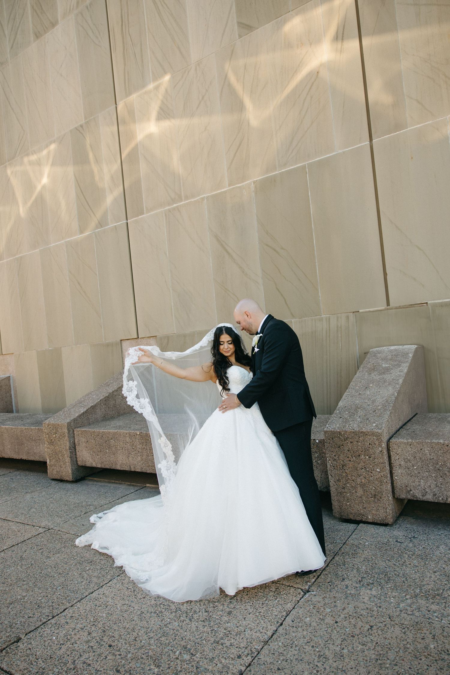 Bride in white ballgown and veil poses with groom in black tuxedo outside modern building on wedding day.