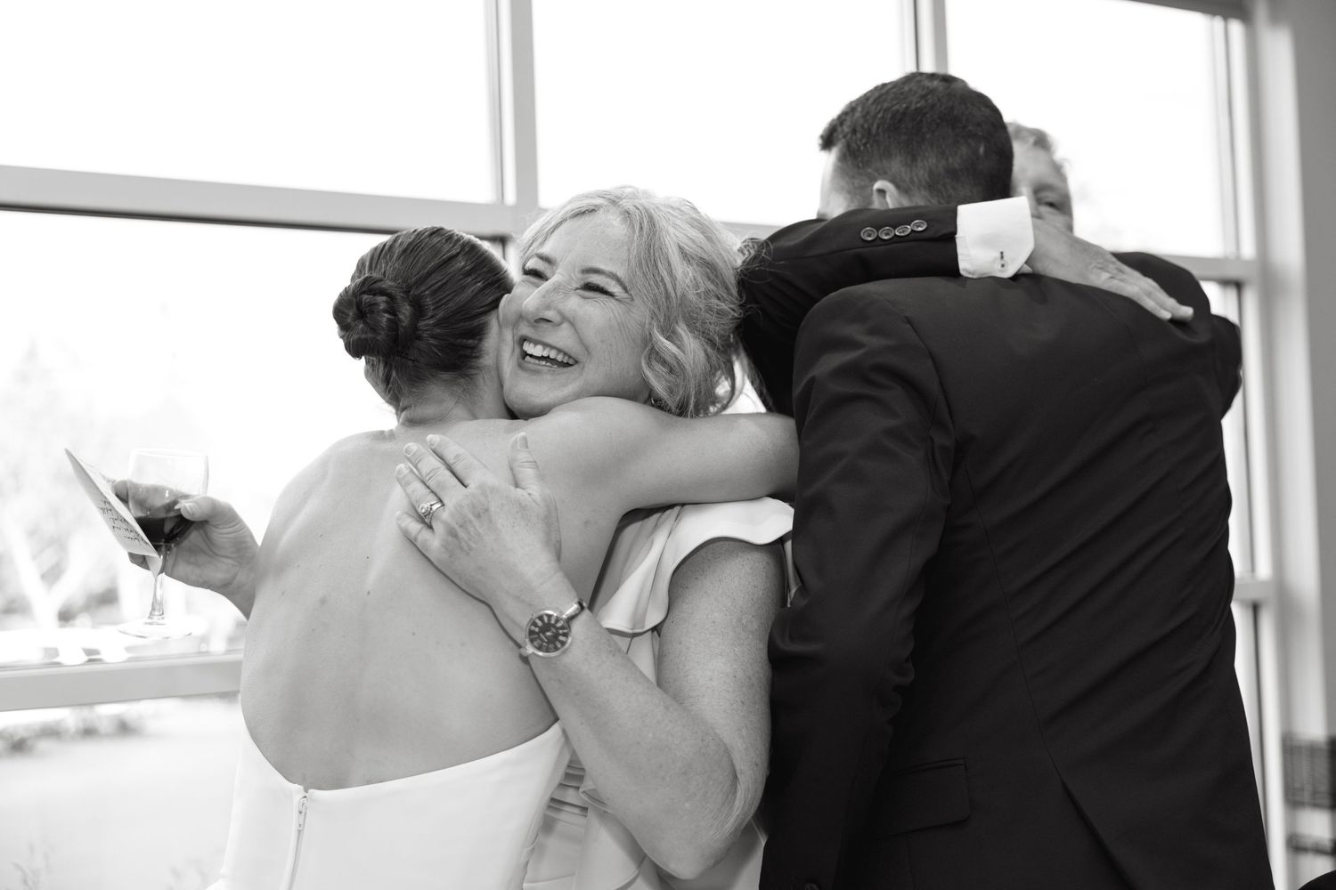 Black and white photo of a bride and groom sharing a warm group hug with a smiling woman at a wedding.