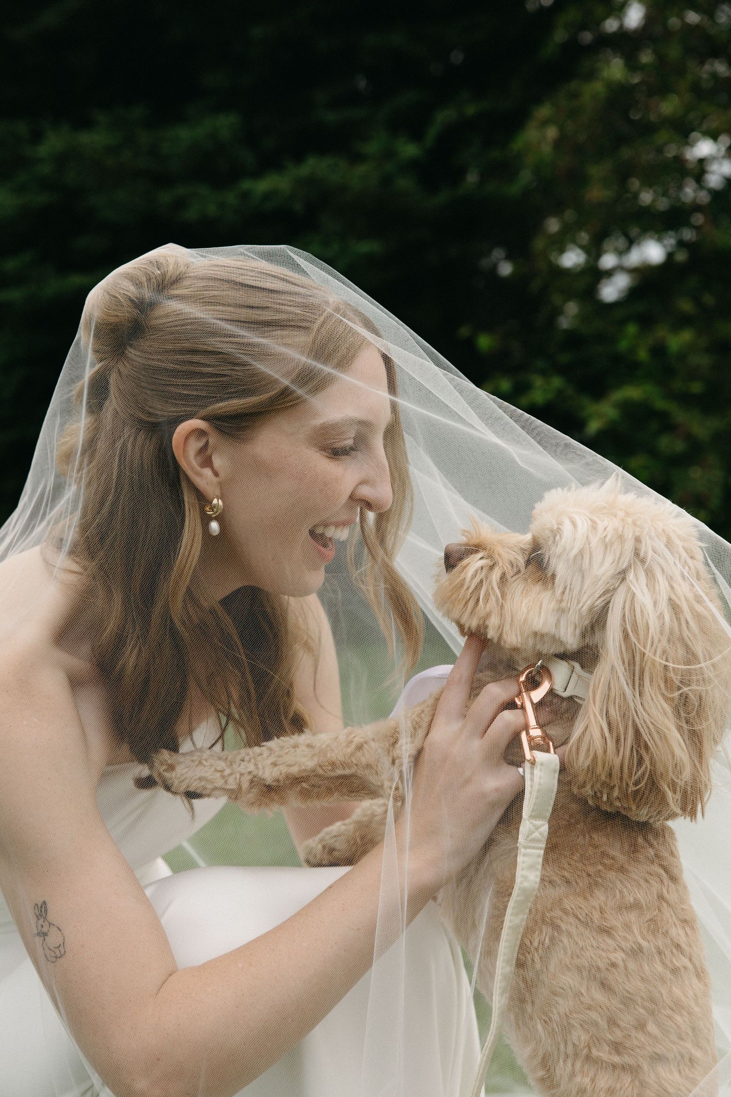 Joyful bride in veil laughing with her fluffy golden doodle dog outdoors on wedding day.