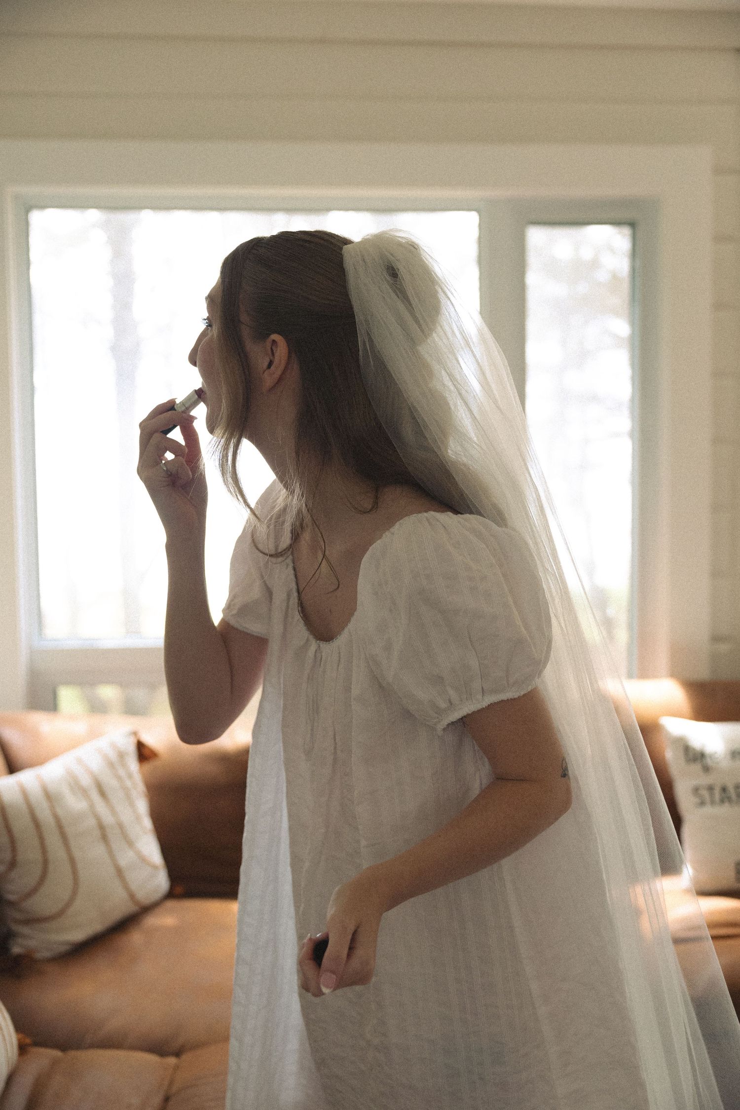 Bride in white dress and veil applying lipstick by window on wedding morning.
