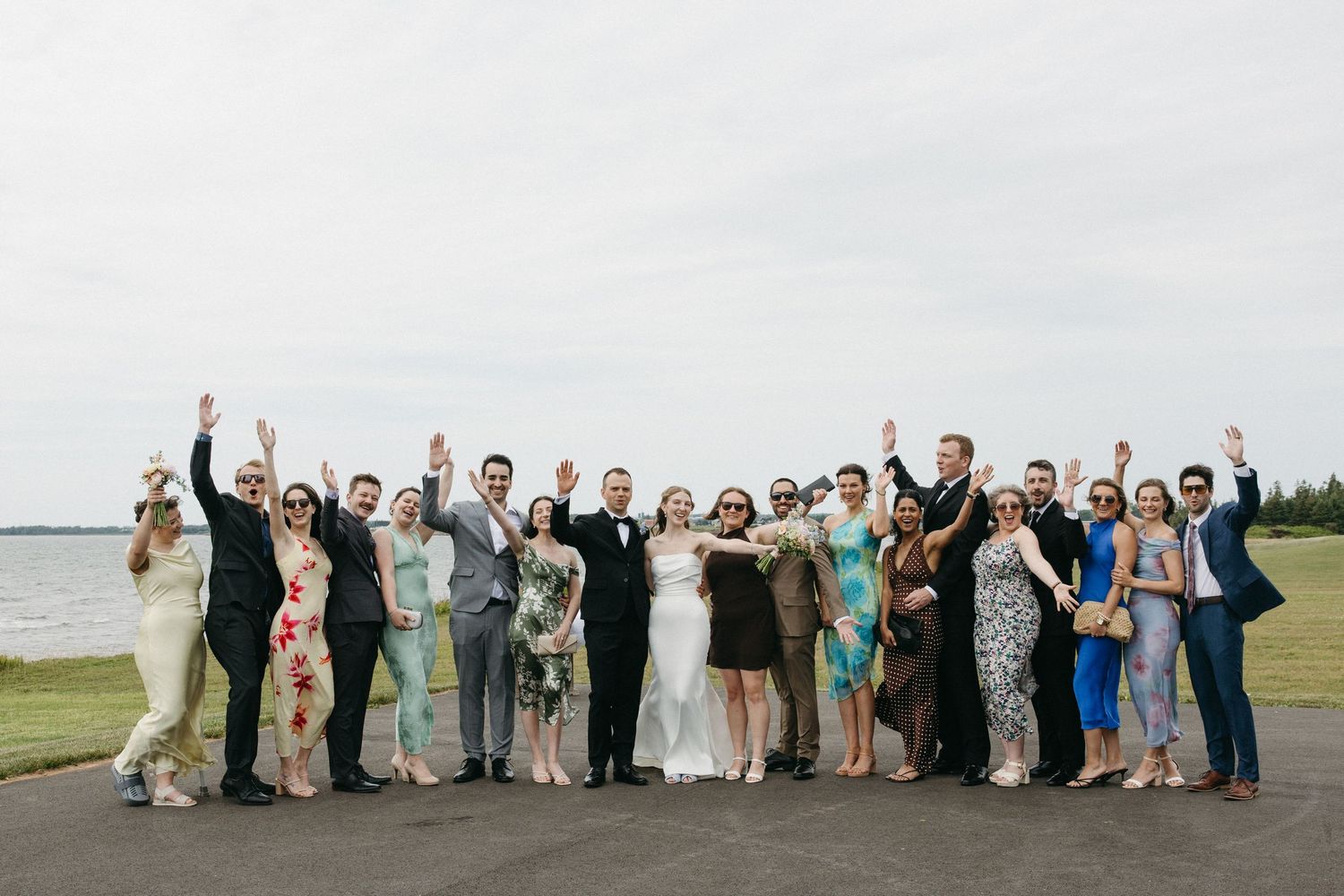 Large wedding party group photo with raised hands celebrating near a waterfront under an overcast sky.