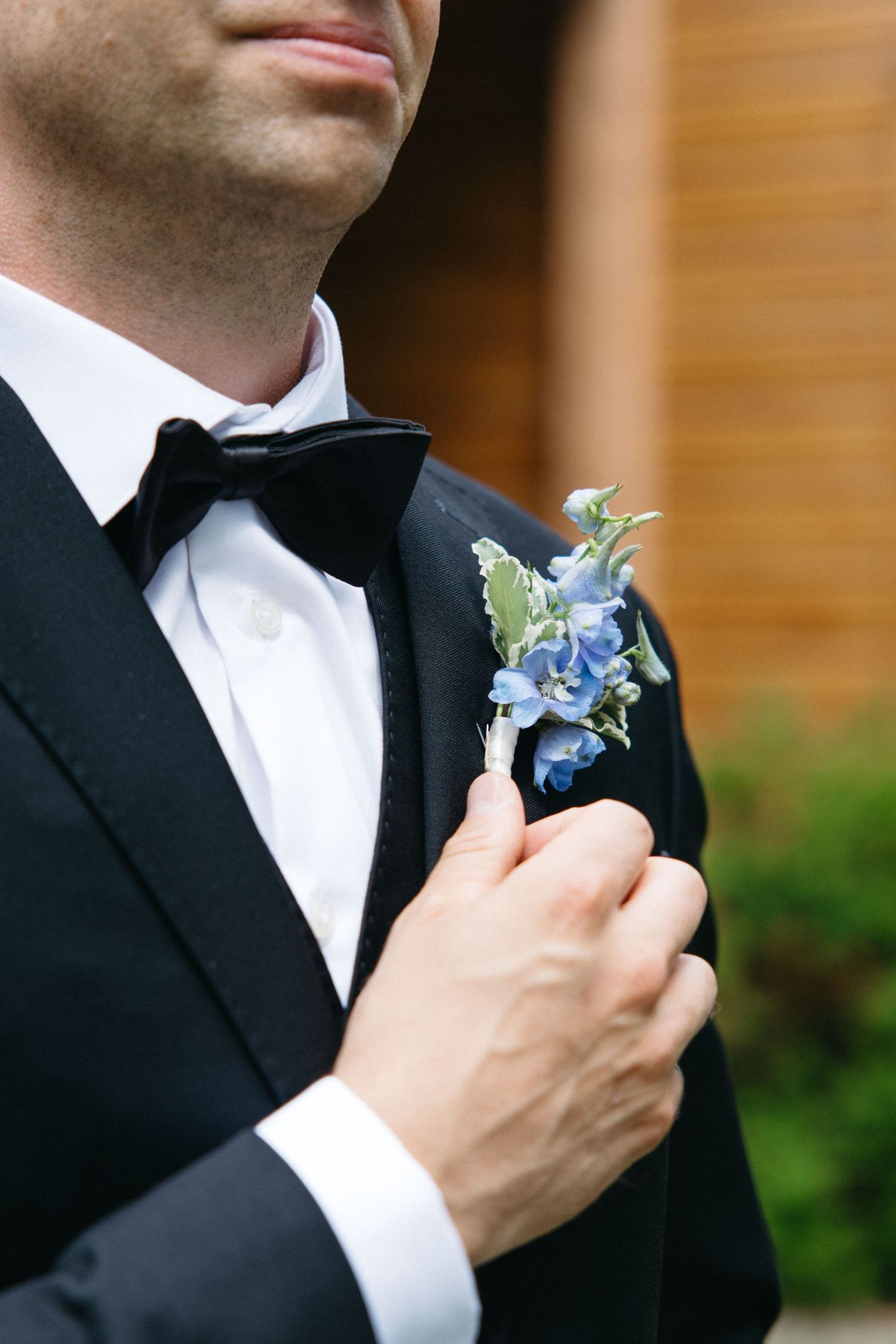Groom in black tuxedo adjusting blue floral boutonniere on lapel.