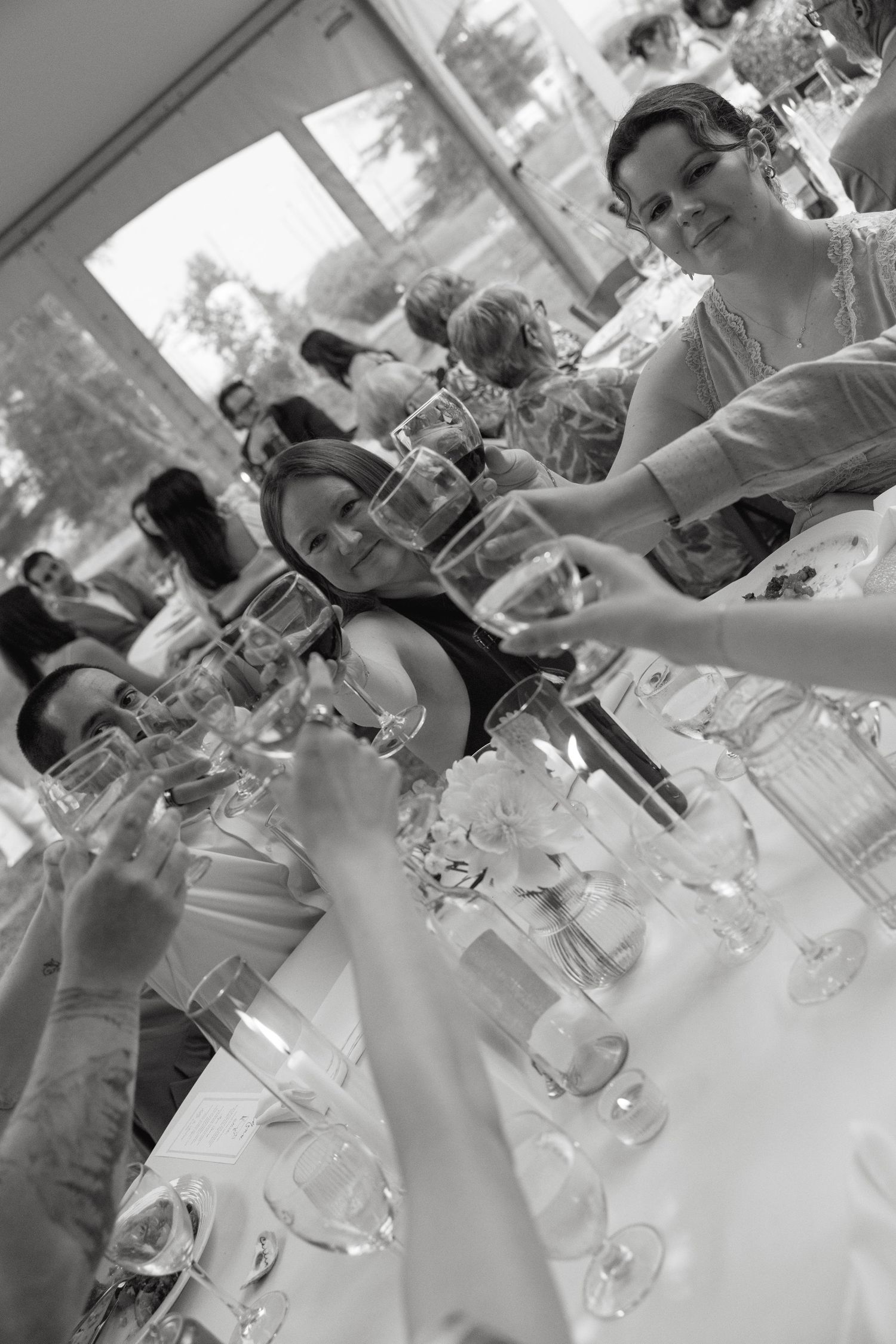 Black and white photo of wedding guests raising champagne glasses in a toast at a decorated reception table.