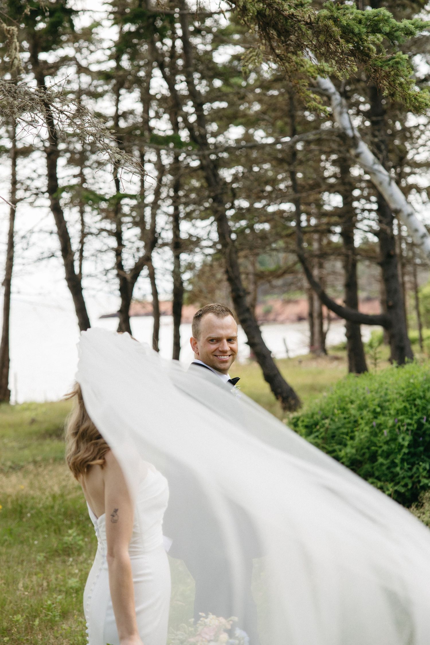 Smiling groom in black tuxedo watches bride's veil billow in the wind among tall pine trees.