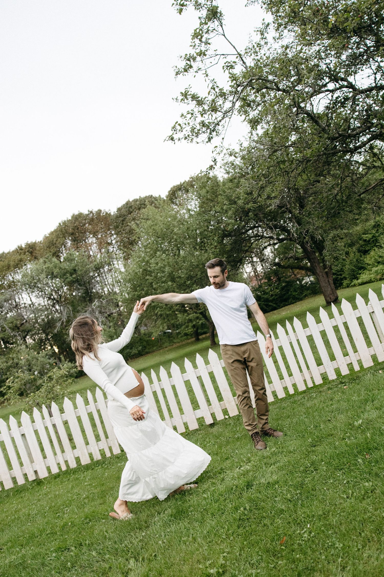 Happy couple in white outfits dance and spin together on green grass near a white picket fence outdoors.