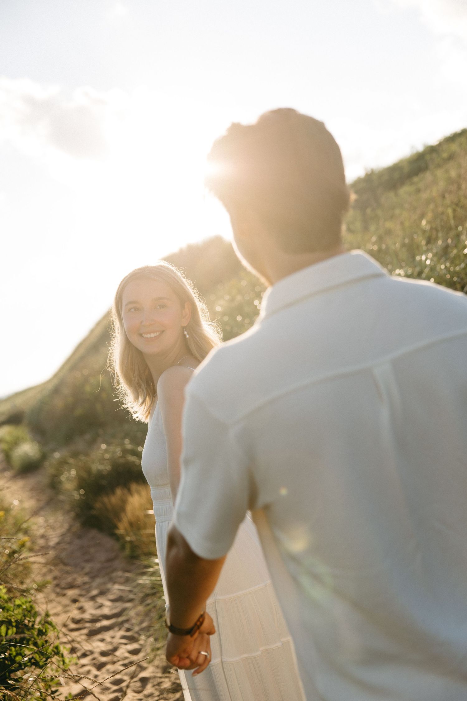 Couple holding hands on sunny beach path, woman smiling back at camera.