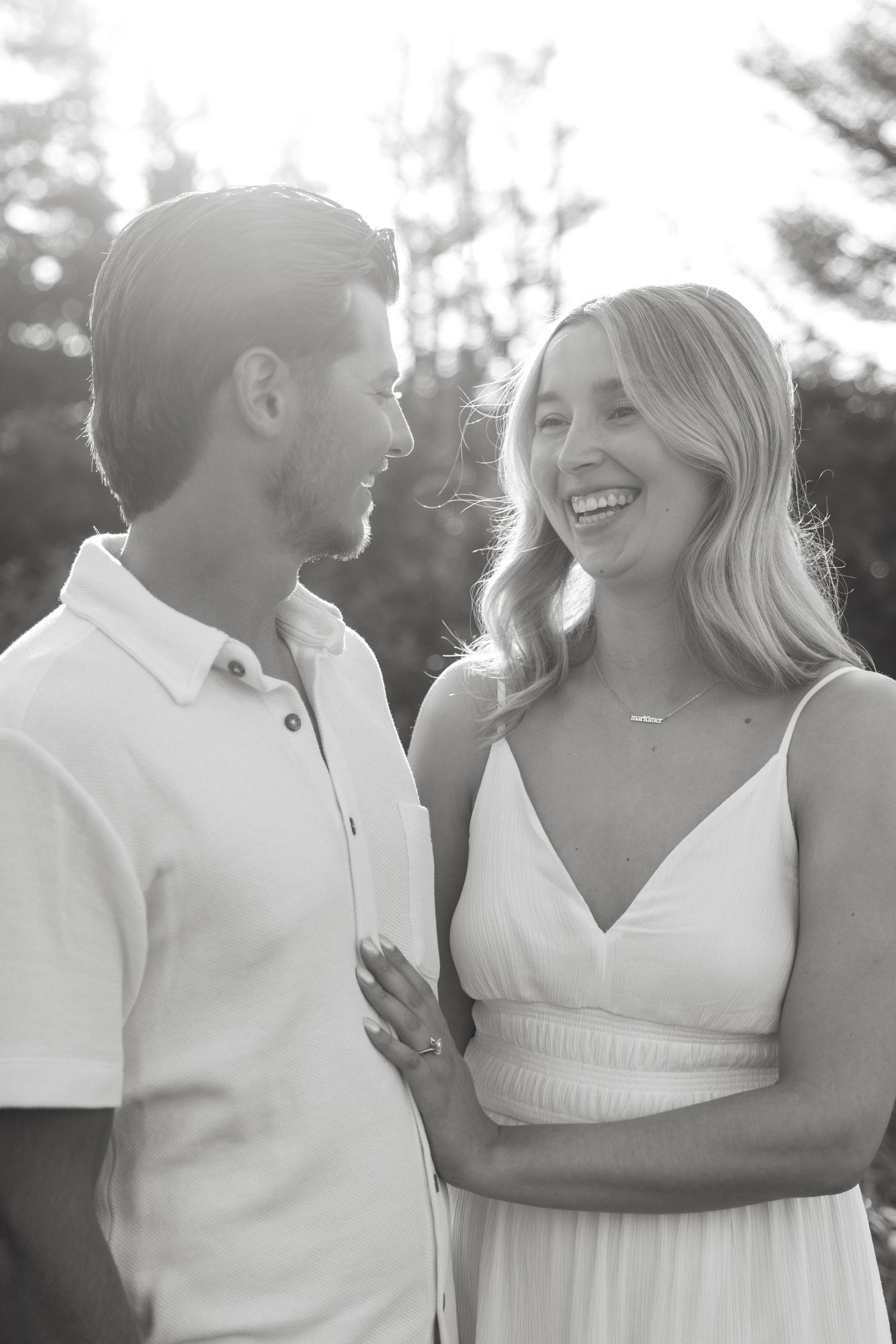 Black and white photo of a smiling couple in white outfits, standing close together outdoors with trees in background.