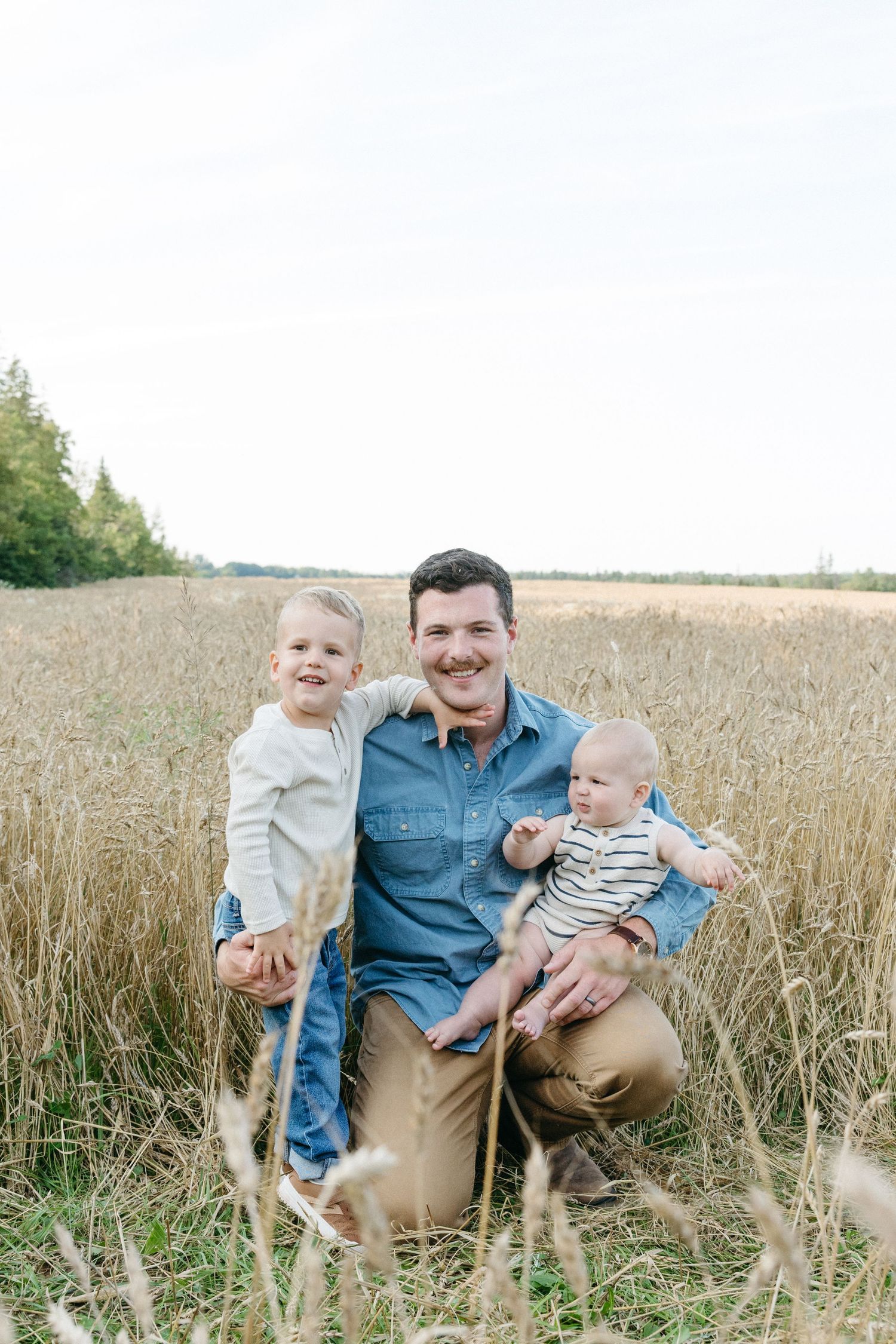 A smiling father crouches in a golden wheat field holding a baby while a toddler stands beside him outdoors.