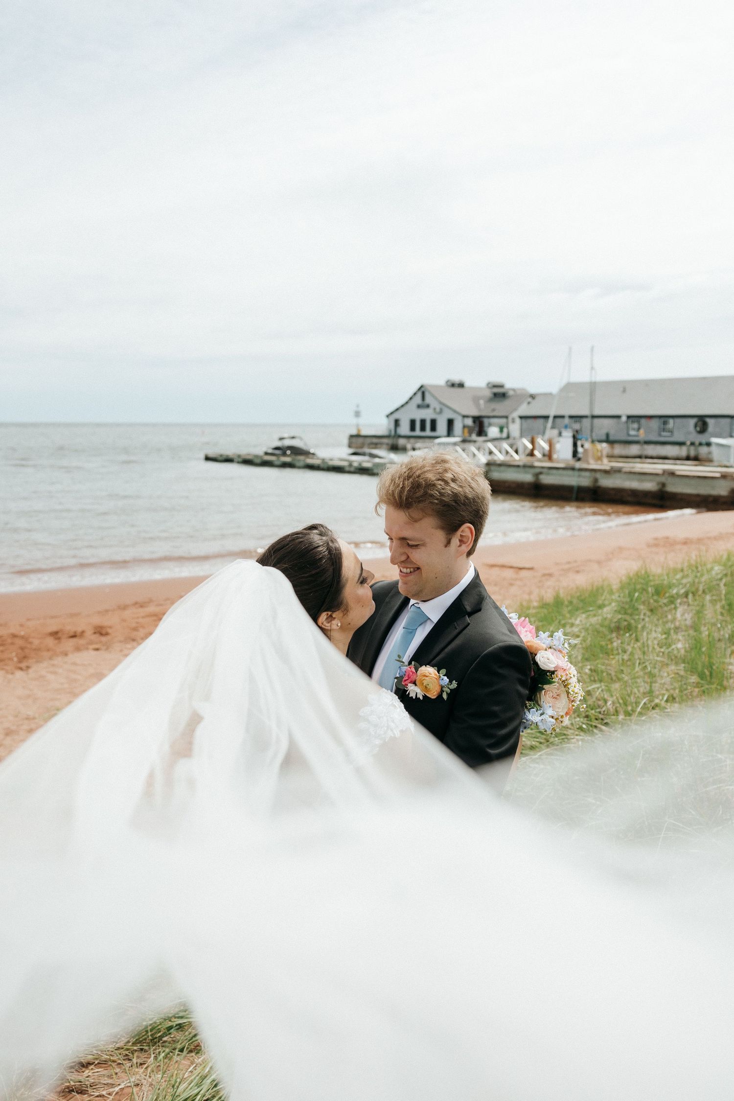A romantic wedding couple shares a kiss by the ocean as the bride's veil flows dramatically in the wind.