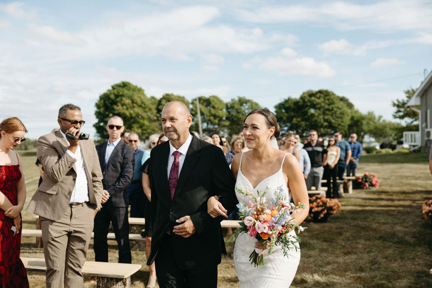 Wedding guests gather outdoors on a sunny day with trees and blue sky in the background.