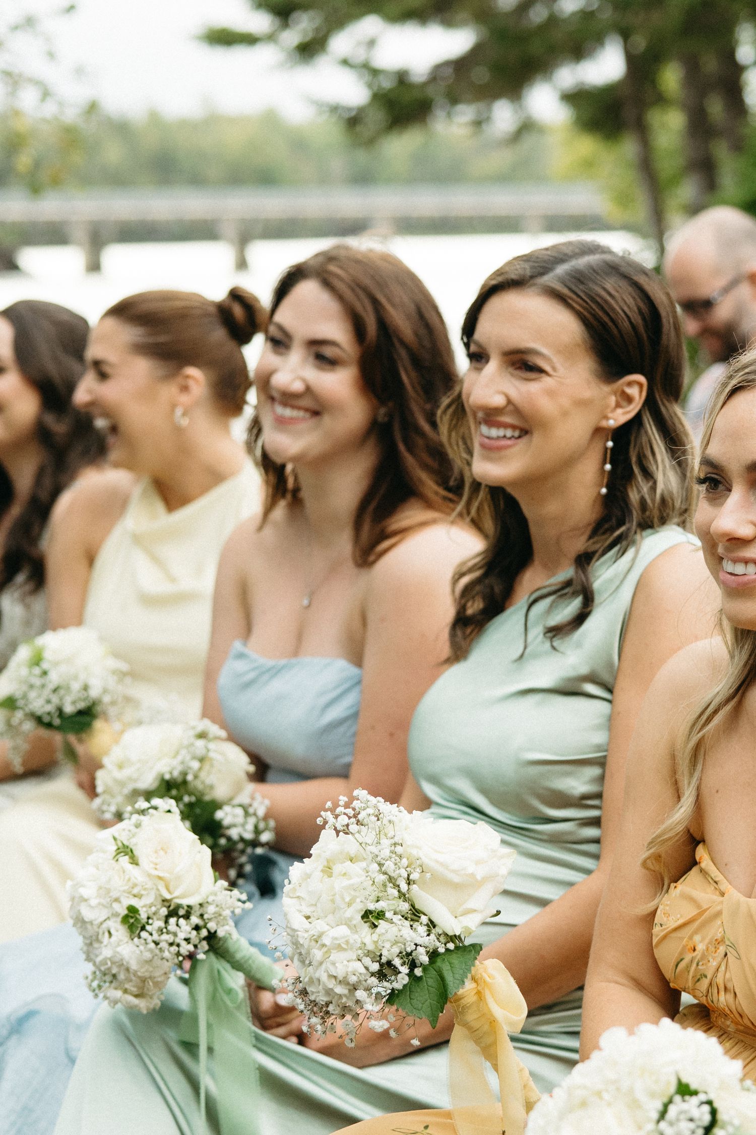 Wedding party members seated outdoors holding white baby's breath bouquets in pastel dresses.