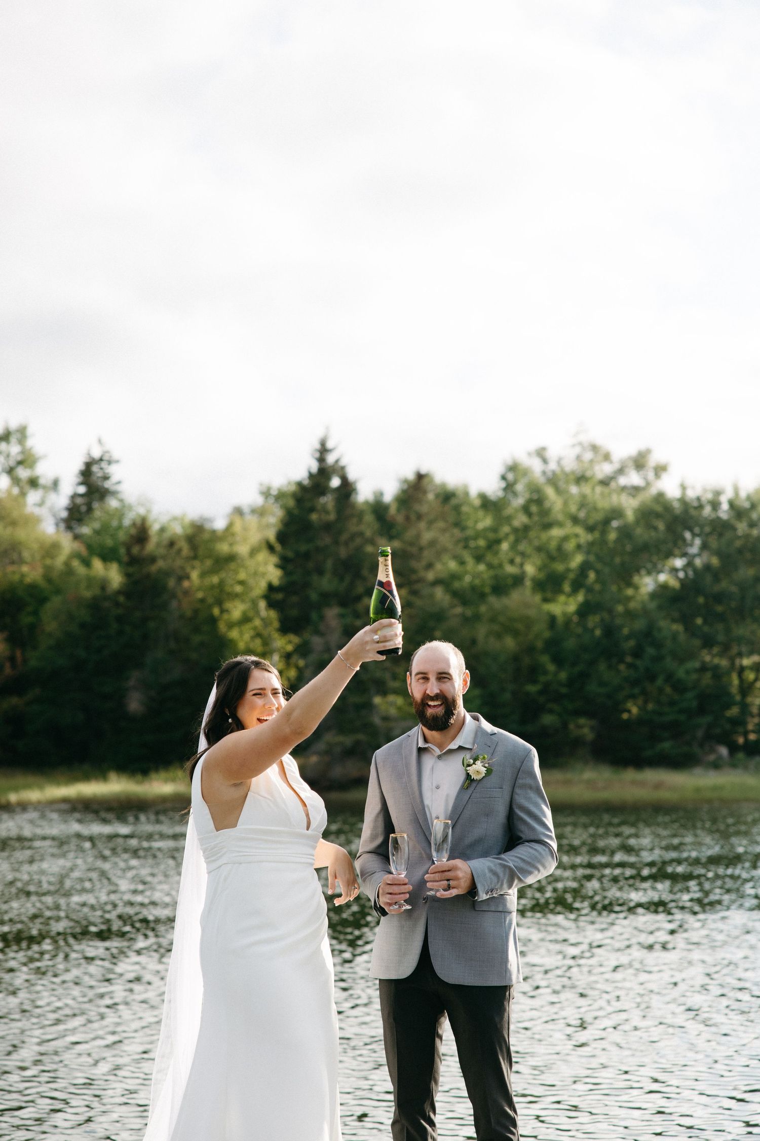 Newlyweds toast with champagne bottles by a serene lake.