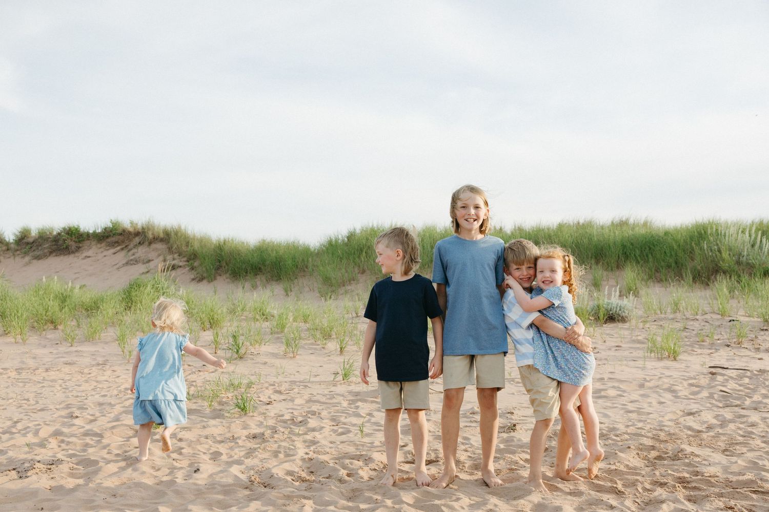 Group of five children standing and hugging together on a sandy beach.