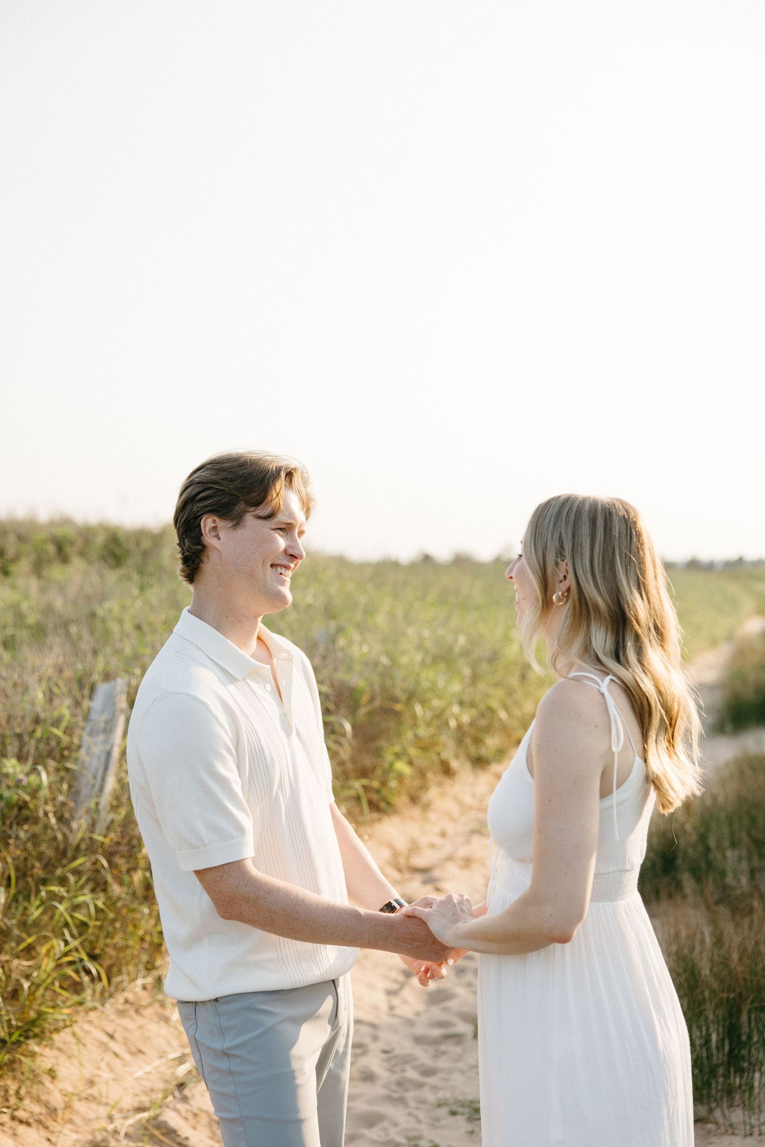 Happy couple holding hands on sandy beach trail surrounded by green dunes in warm sunlight.
