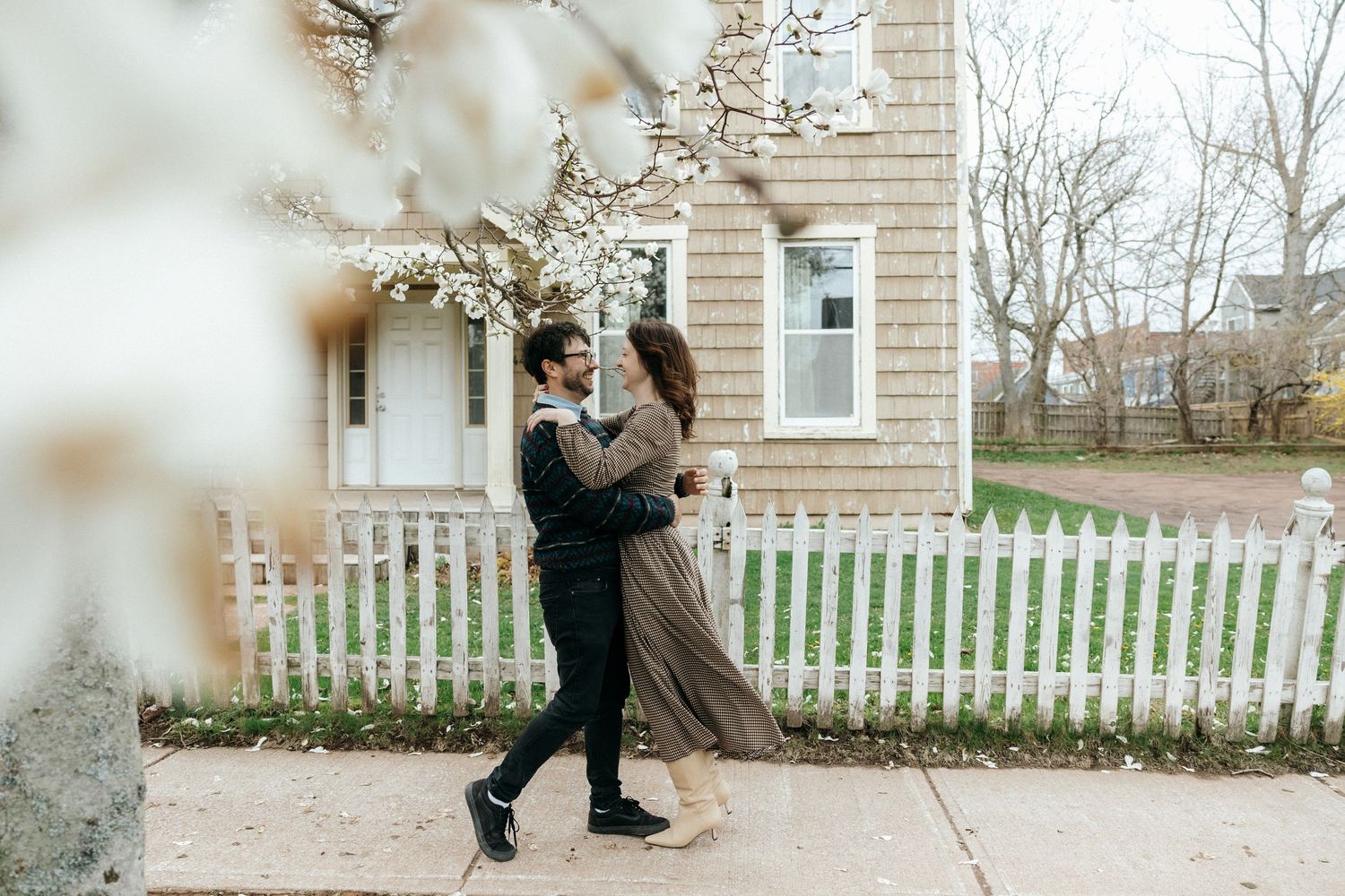 A couple embraces and dances on a sidewalk in front of a house with a white picket fence in spring.