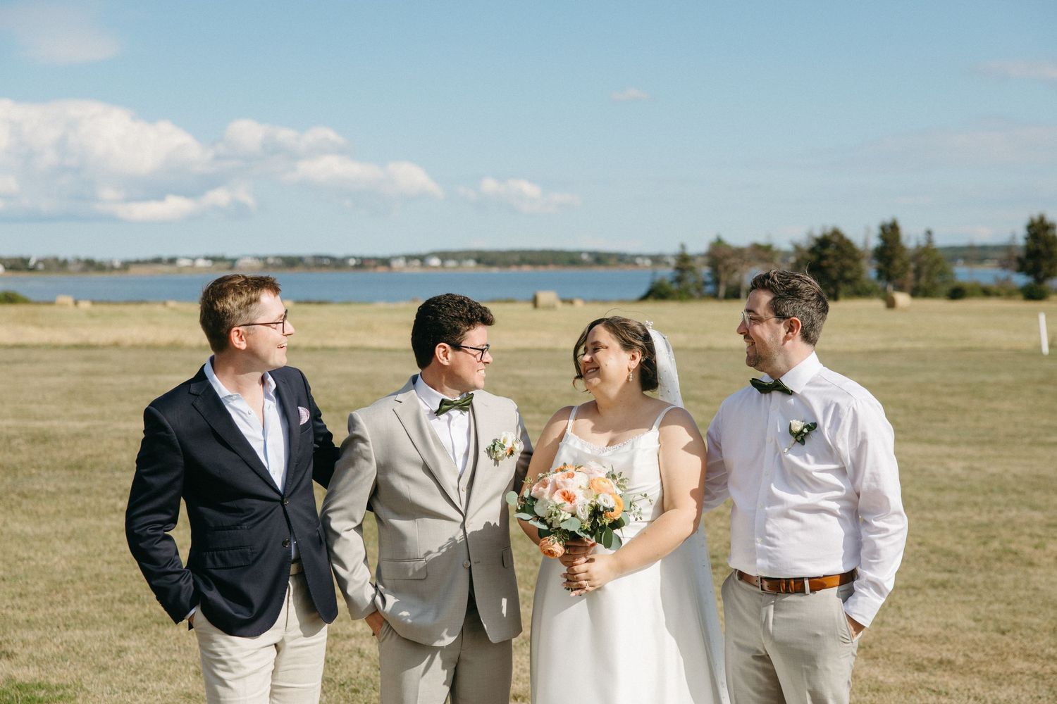 Groom in gray suit with bow tie stands with bride and two guests in a sunny waterfront field.