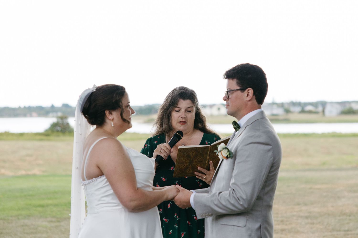 Bride and groom holding hands during wedding ceremony officiated by a woman in a green dress outdoors.