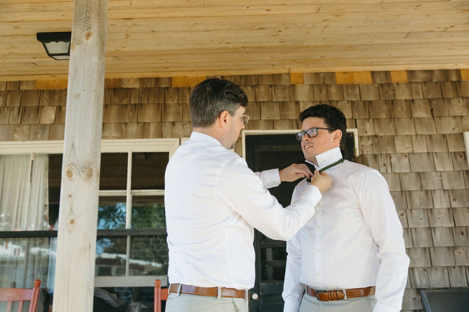Groomsman helping groom adjust bow tie outside a rustic shingle-sided building on a sunny wedding day.
