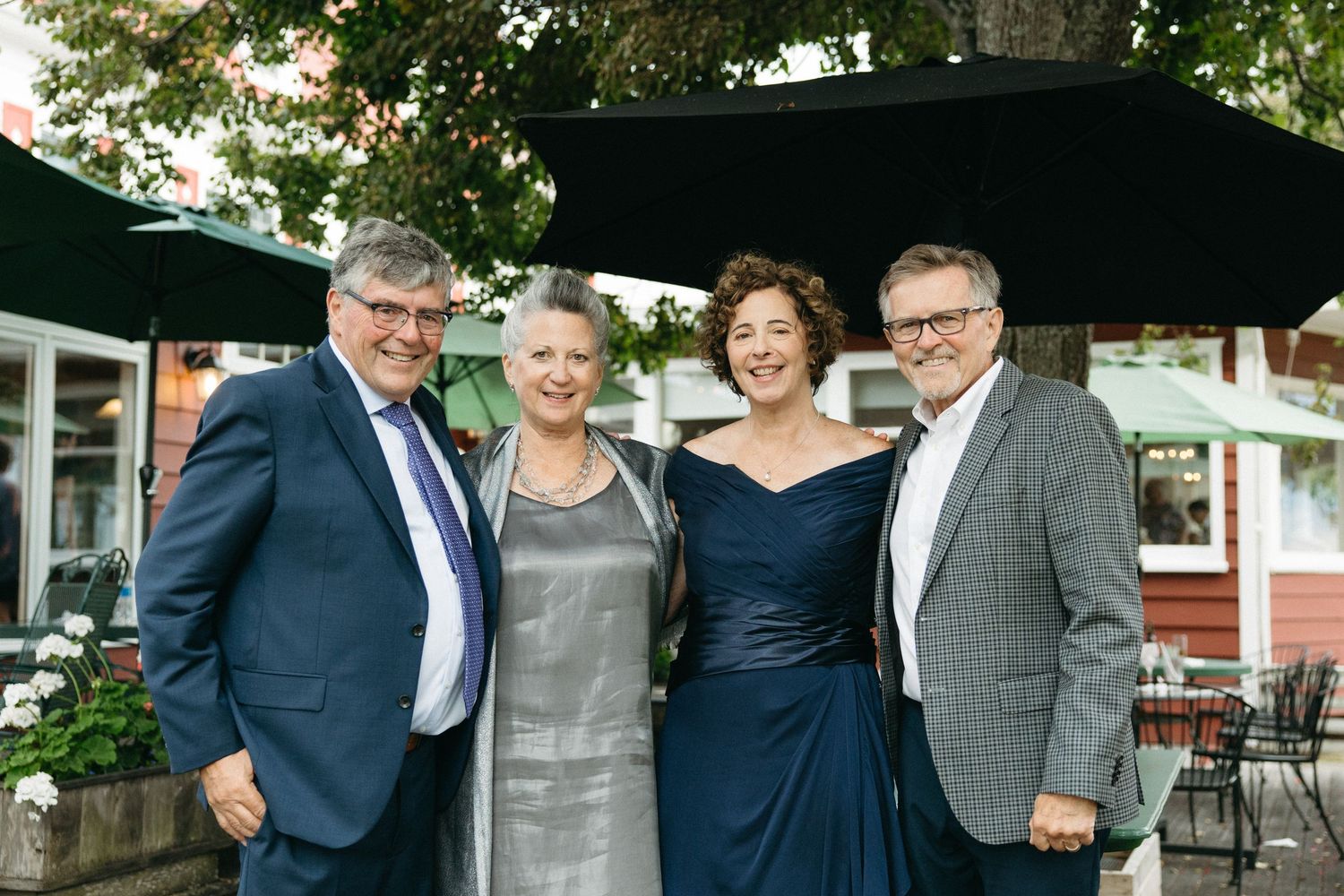 Two couples smile together outdoors at a wedding reception under green umbrellas in evening light.