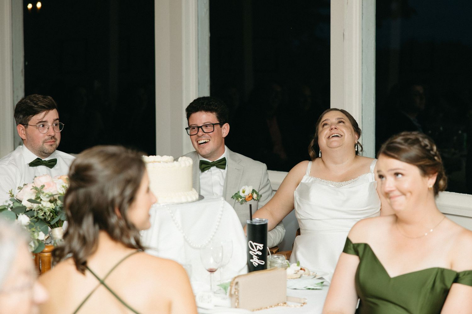 Bride and groom laugh joyfully at wedding reception head table with wedding cake nearby.