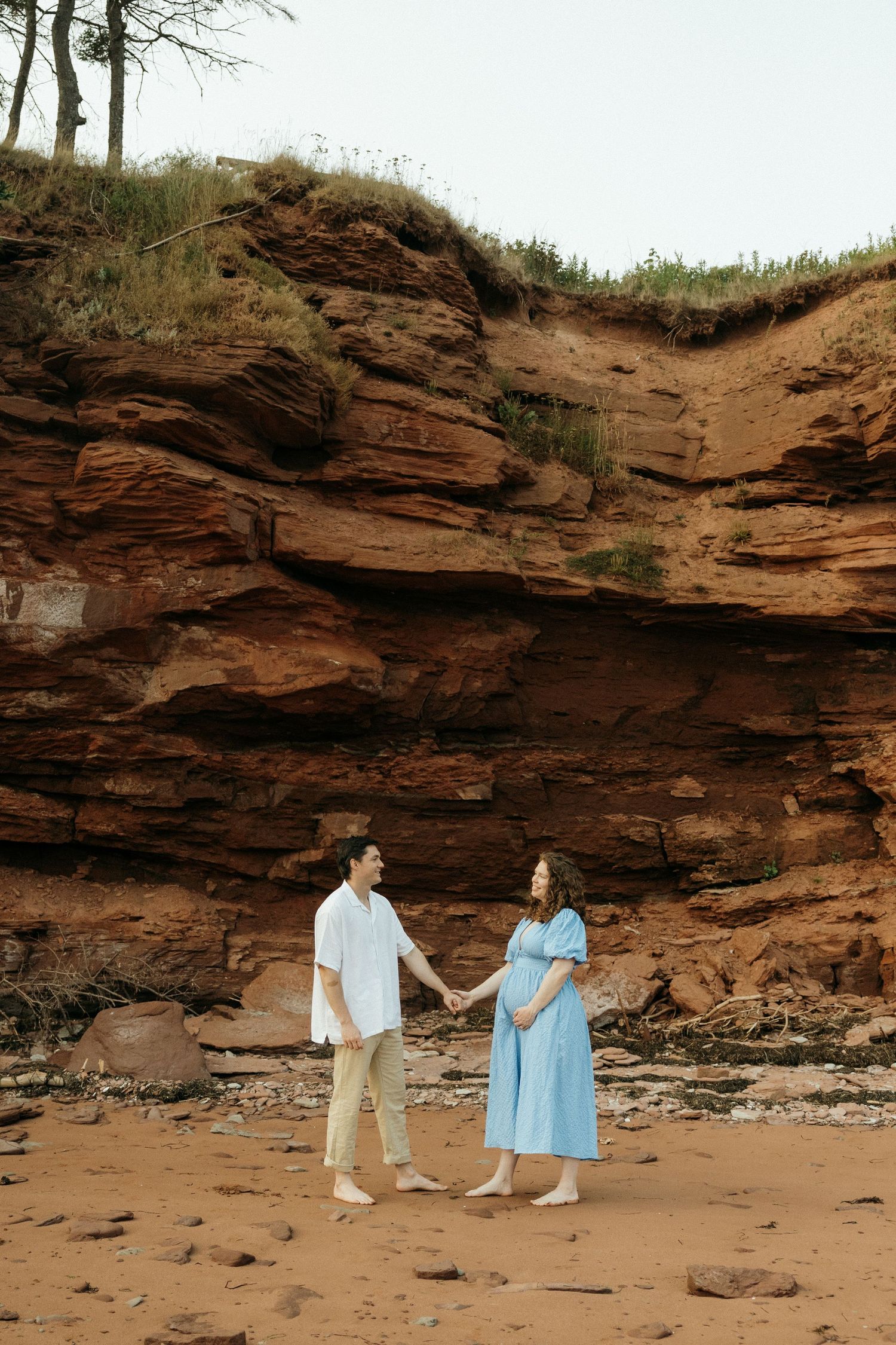 A couple walks hand in hand along a beach with towering red cliffs in the background.