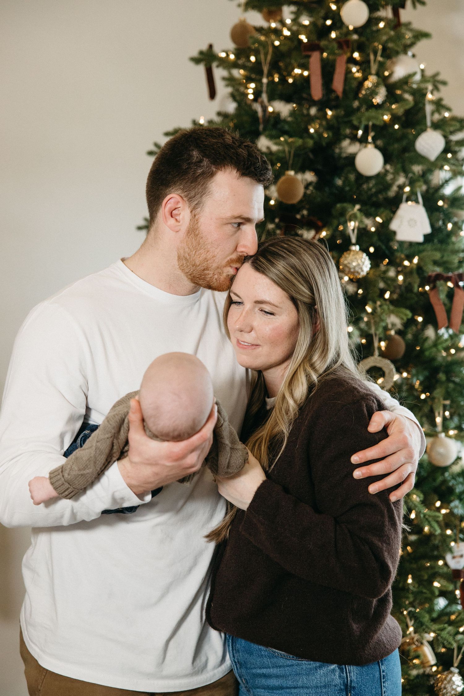 Couple embraces with baby in brown sweater near decorated Christmas tree with warm lighting.