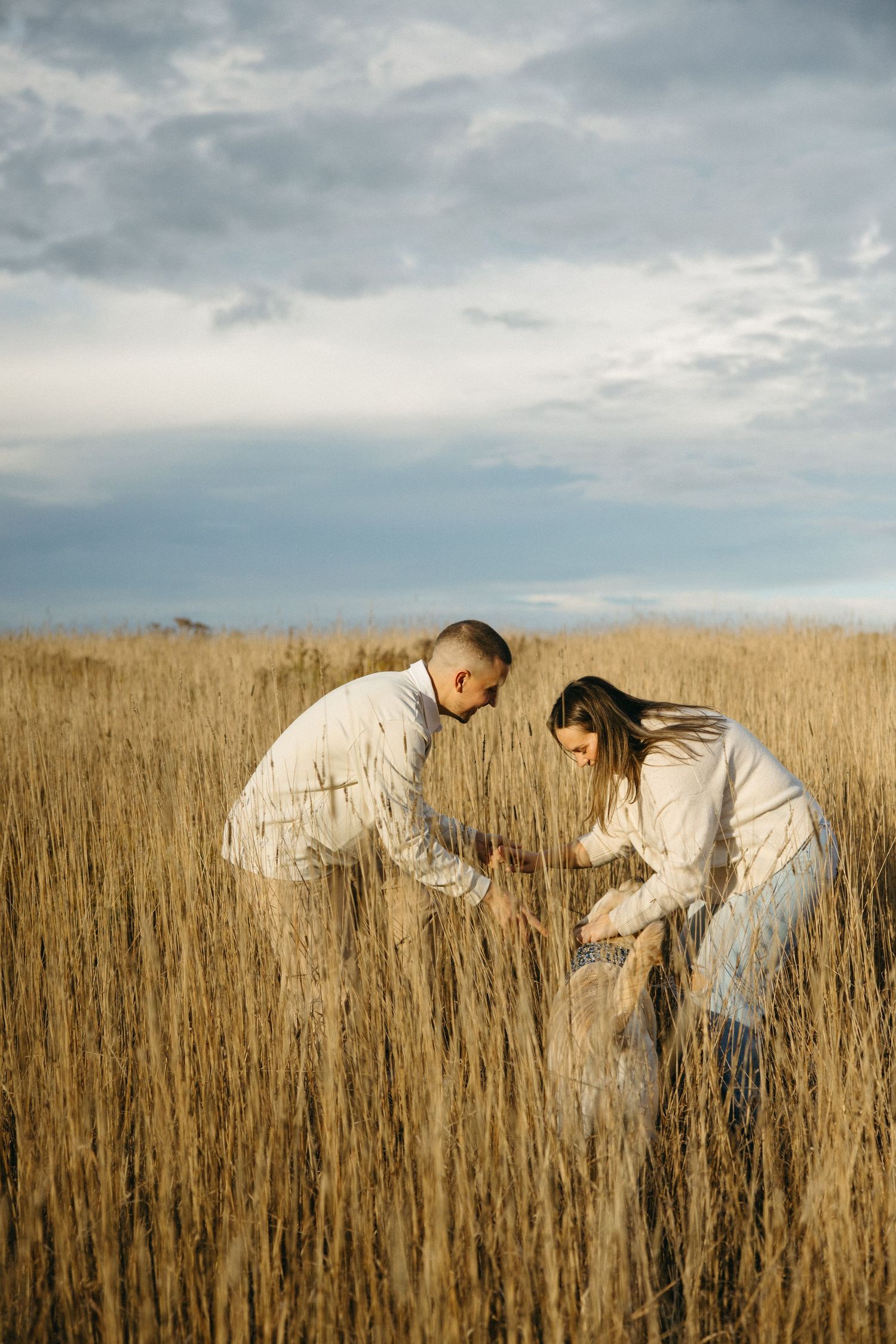 Candid moment of two people bending down in a wheat field under cloudy skies.