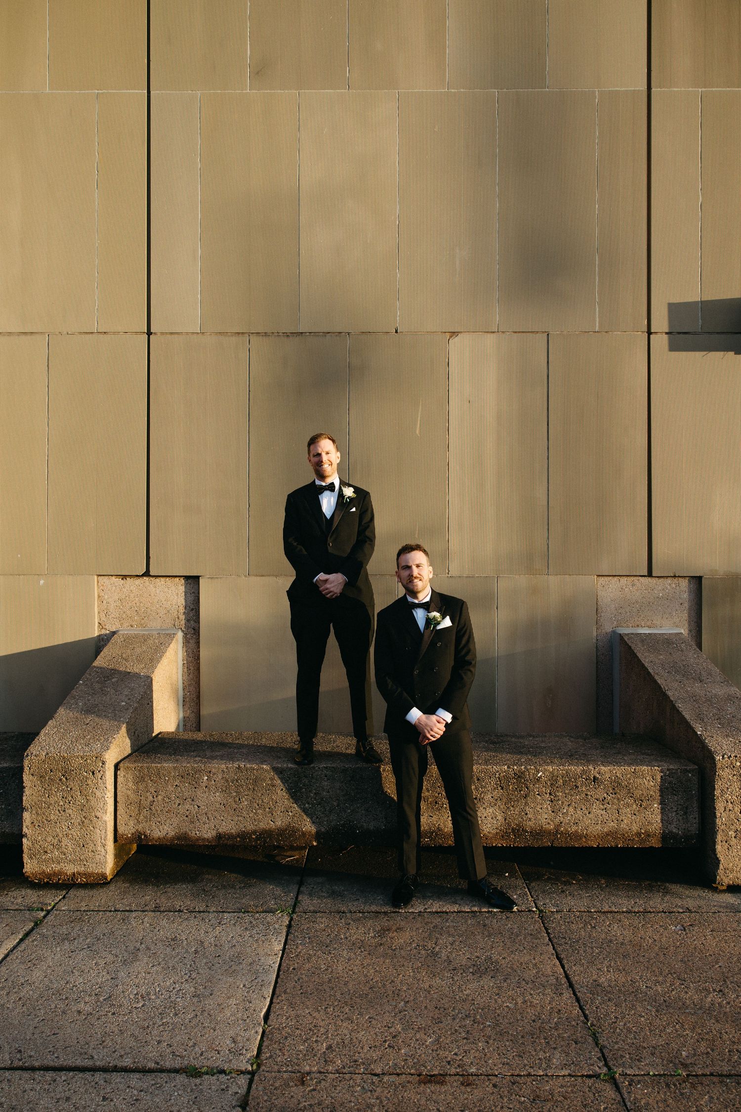 Two people in black tuxedos standing against stone wall in dramatic lighting.