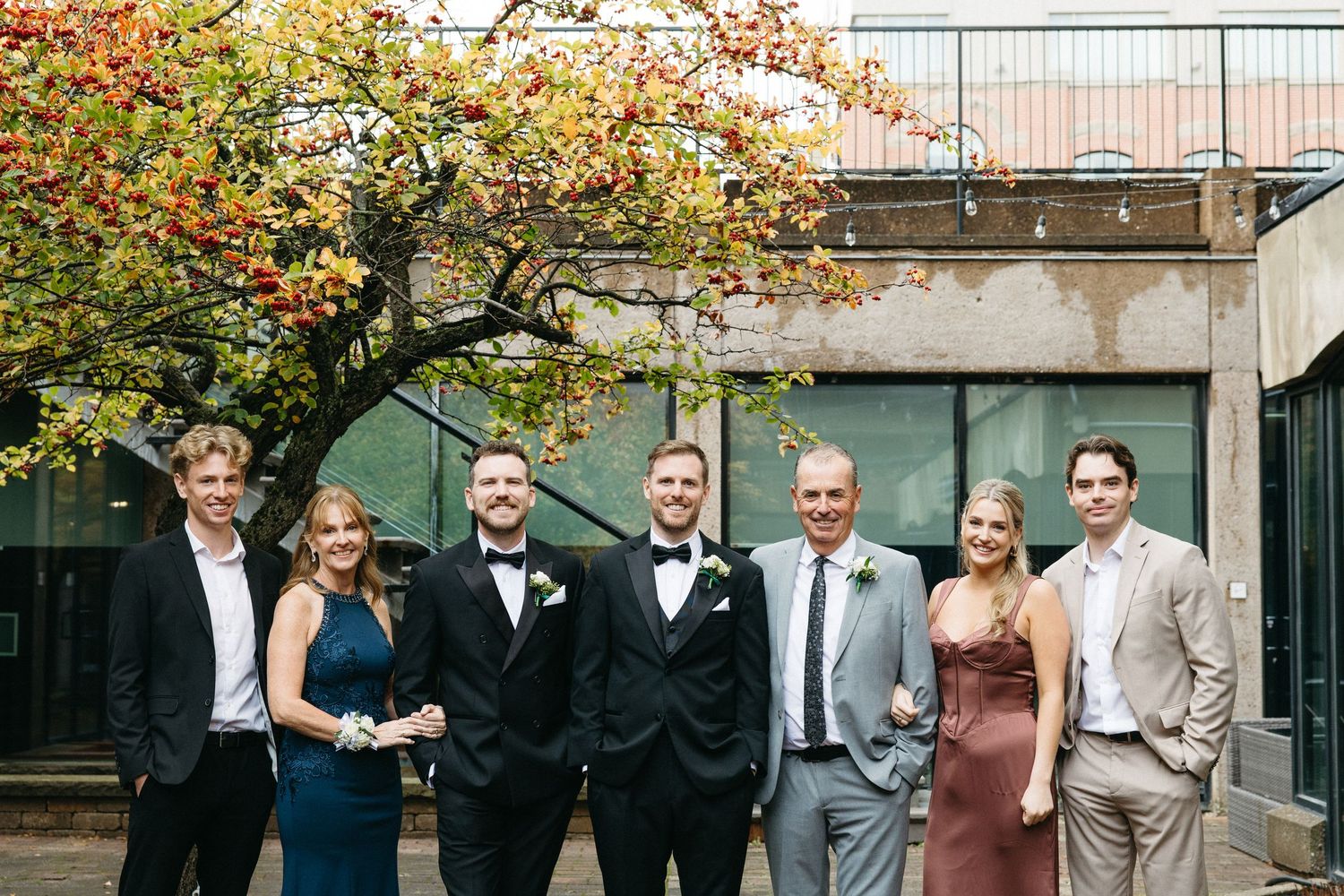 A group in formal evening wear poses together under golden autumn leaves in an outdoor courtyard.