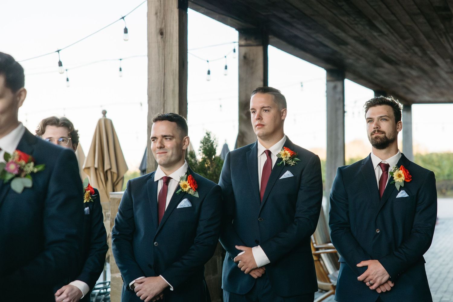 Wedding party members in dark suits and red boutonnieres stand together at an indoor venue.