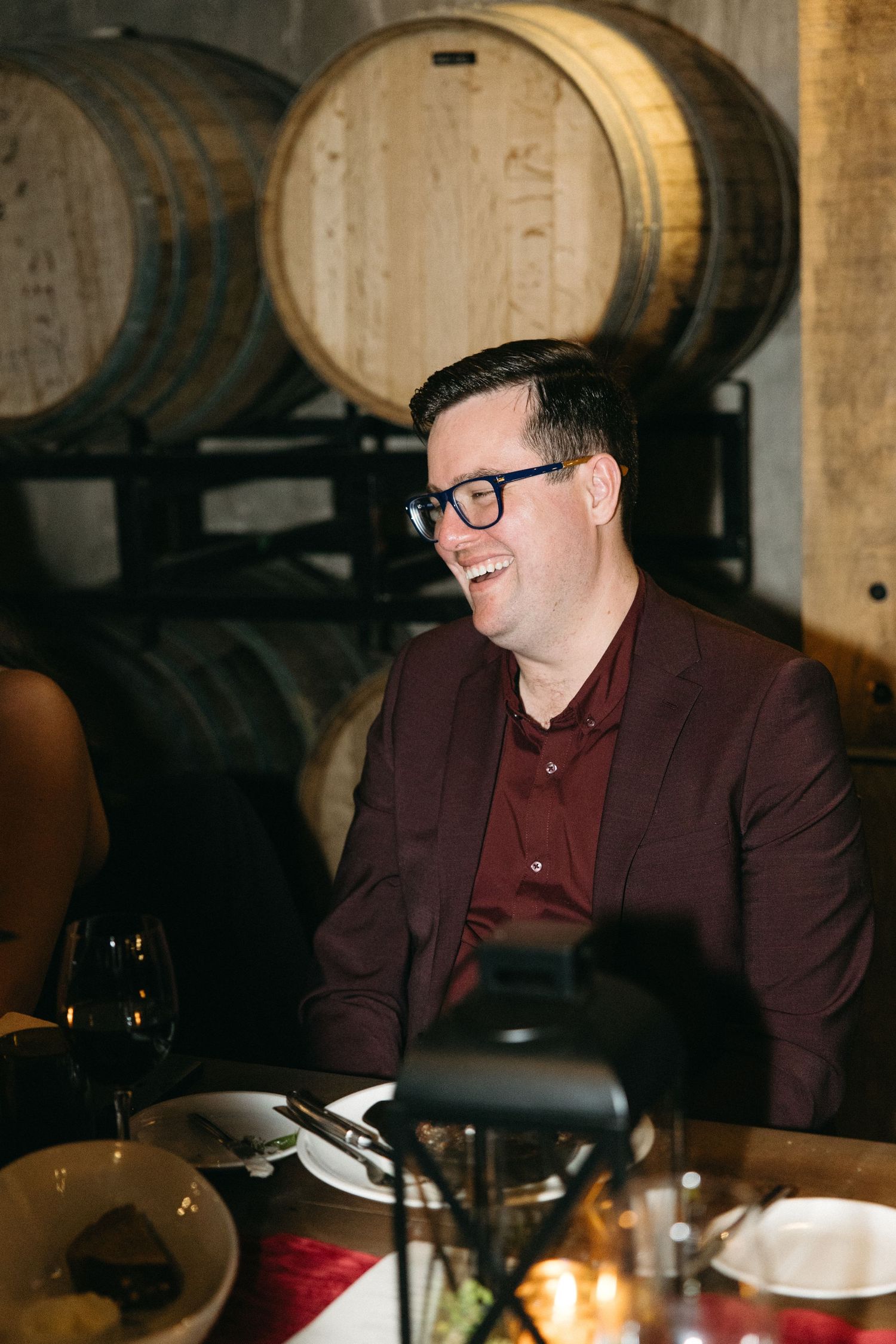 Guest in burgundy suit jacket seated at dinner table with wine barrels visible behind.